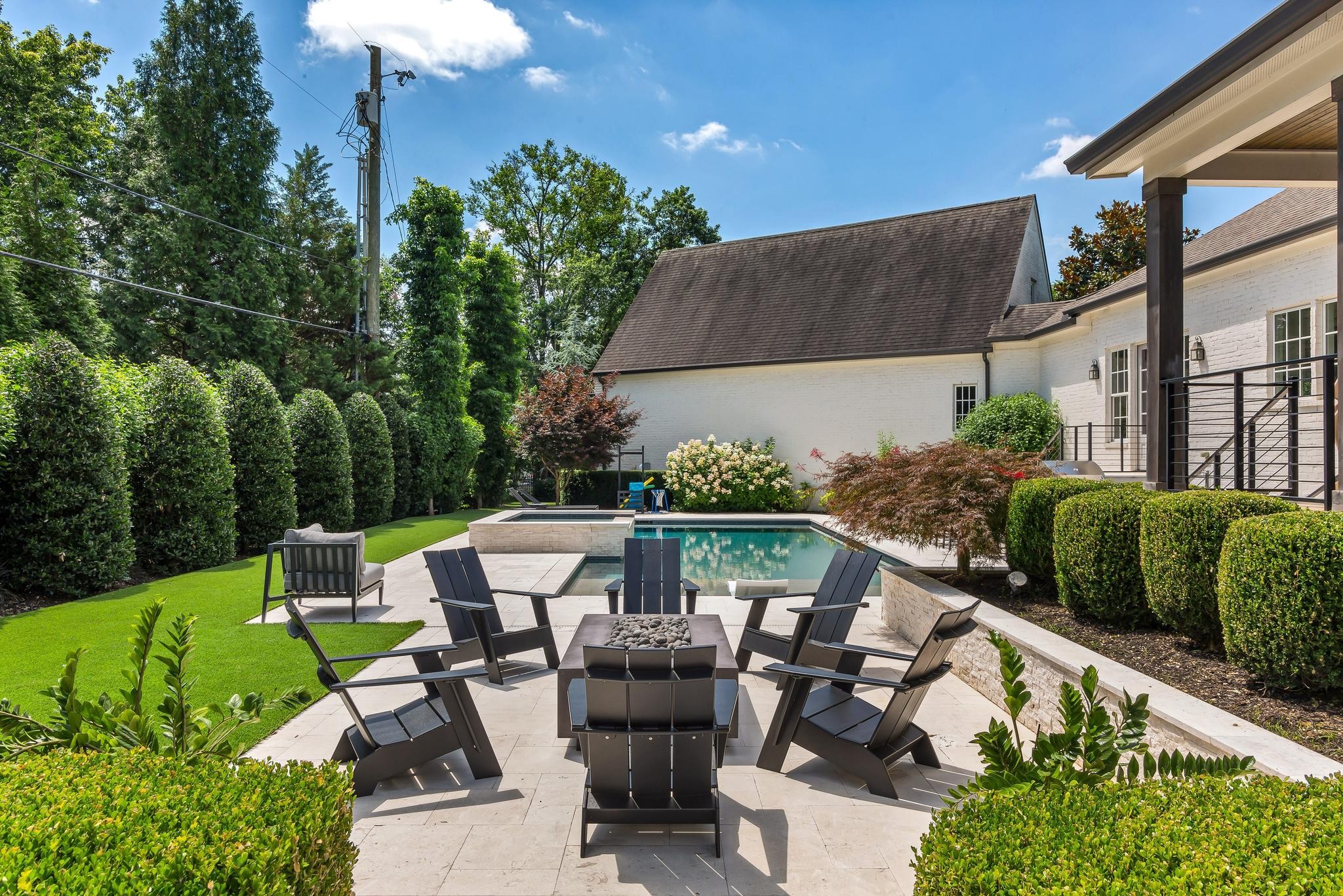 Backyard pool and stone patio with dark lounge chairs, manicured hedges, and a covered porch.