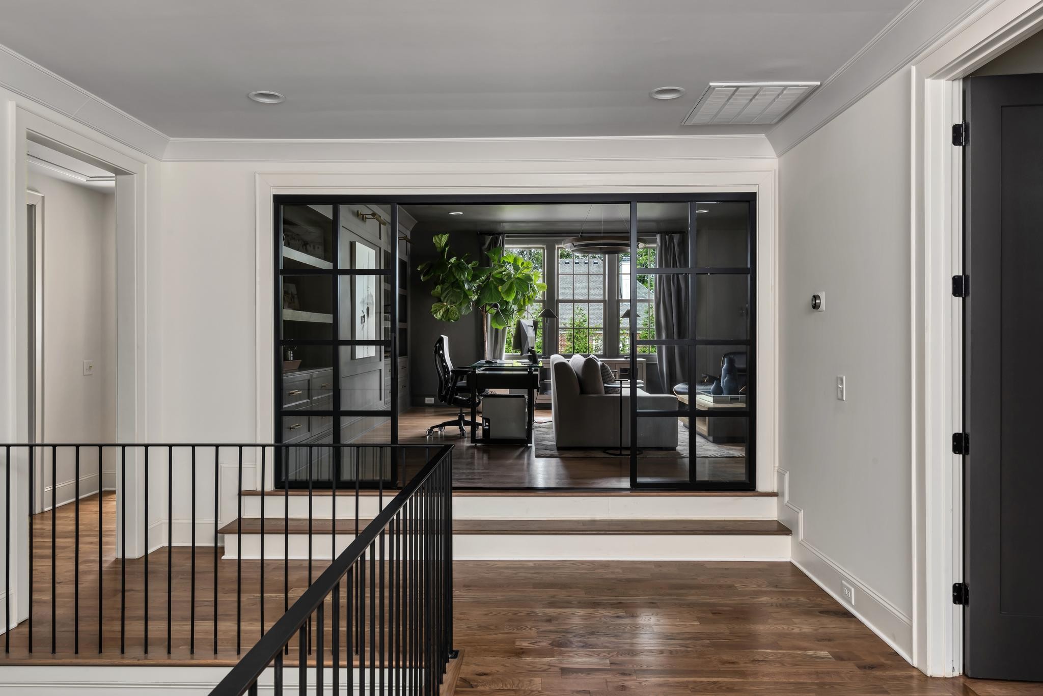 Hardwood landing overlooking a glass-enclosed office with a desk, leafy plants, and gray seating.