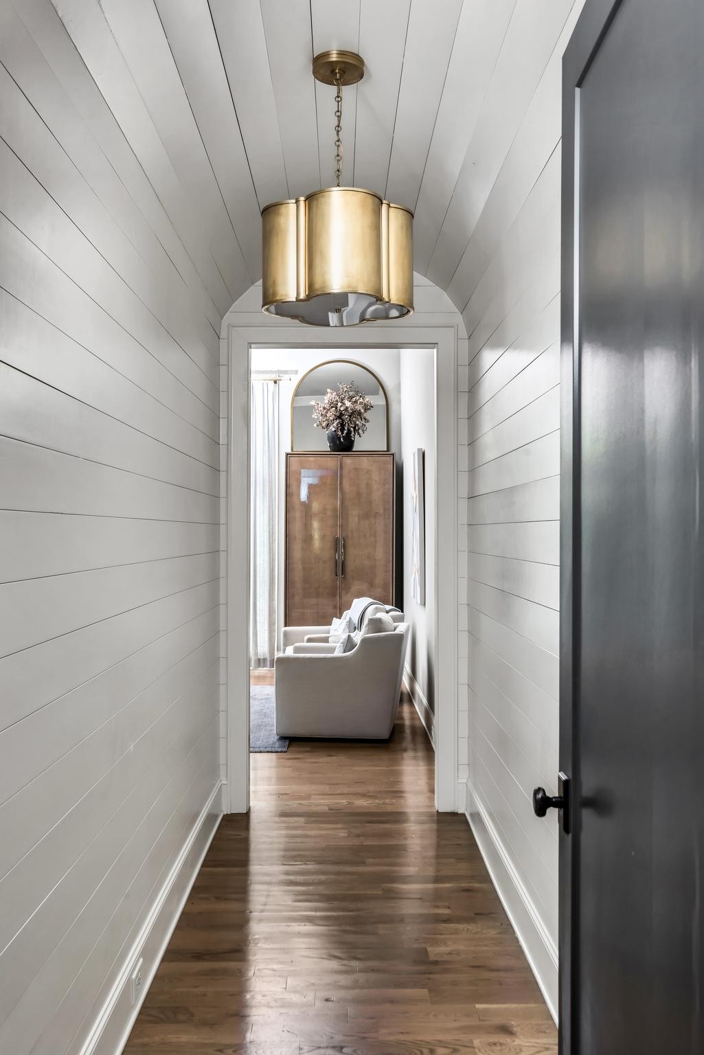 Hallway with white paneled walls, vaulted ceiling, brass pendant, and a sitting room beyond.
