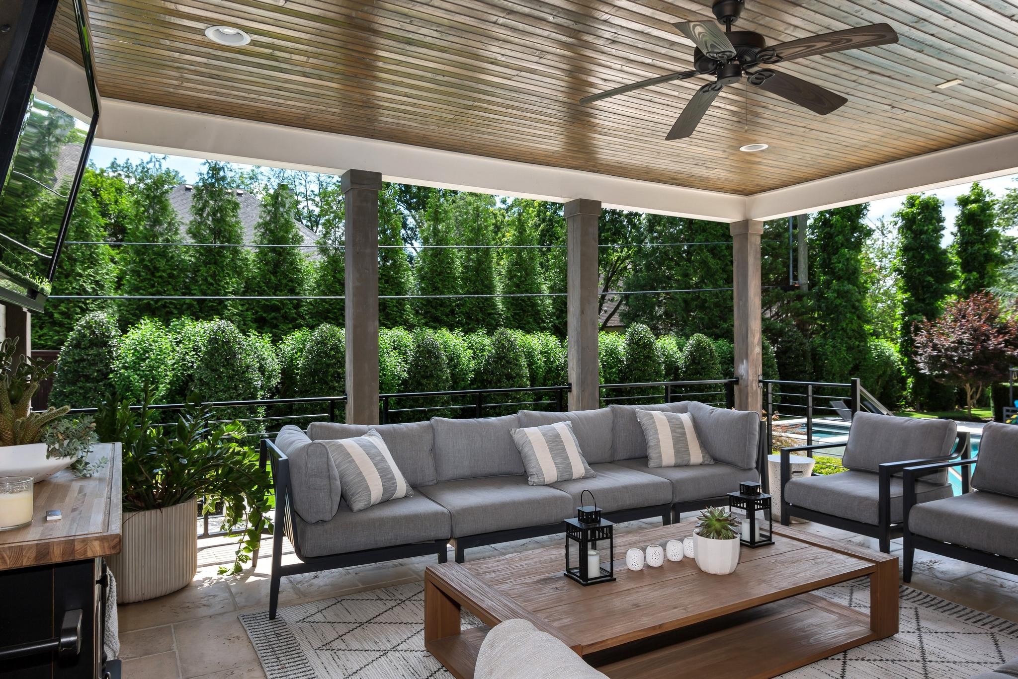 Covered patio with gray lounge seating, a wood-plank ceiling, and views of greenery and a pool.