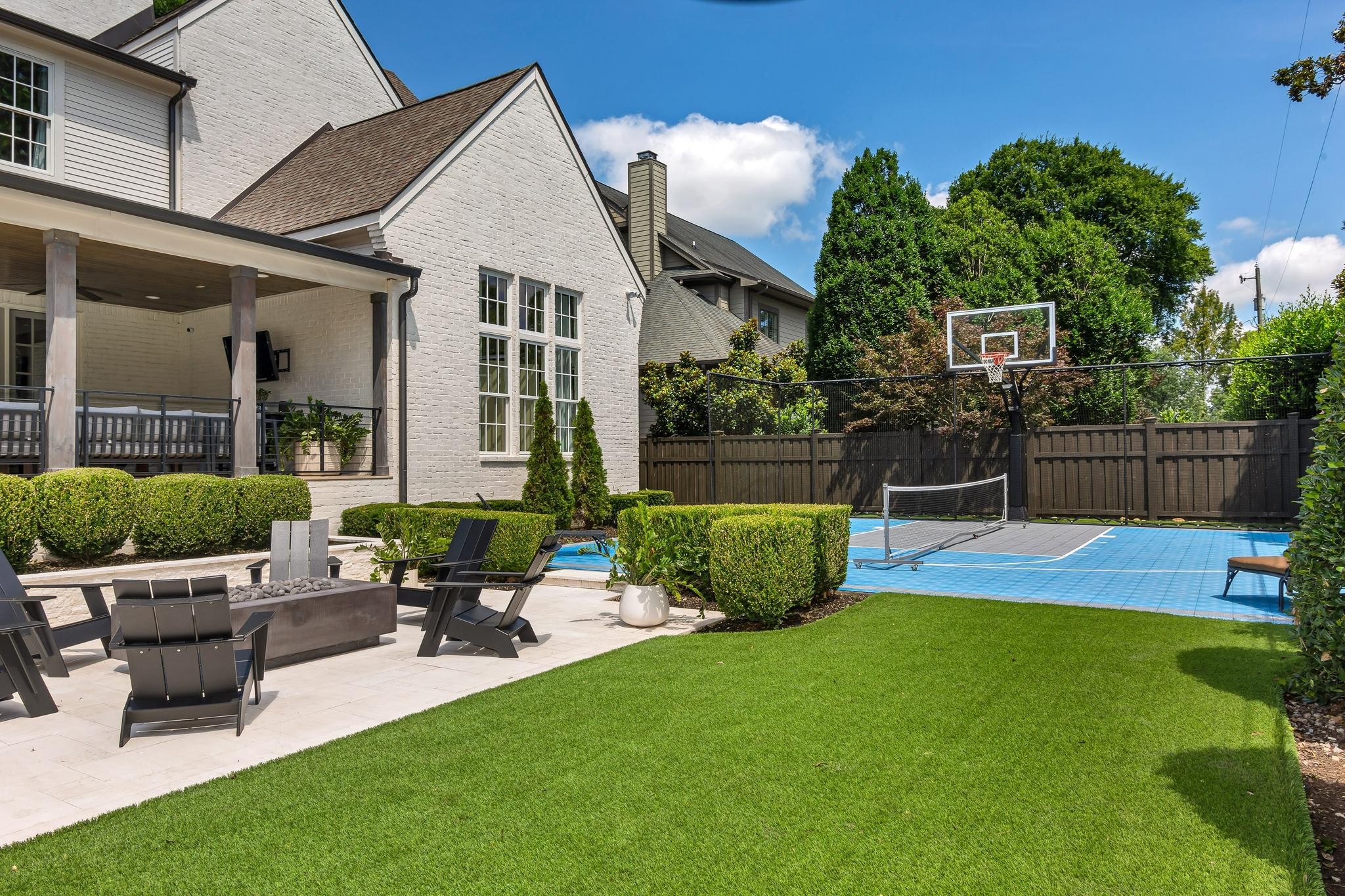 Backyard with a white brick home, covered patio, manicured lawn, and blue sport court with basketball hoop.