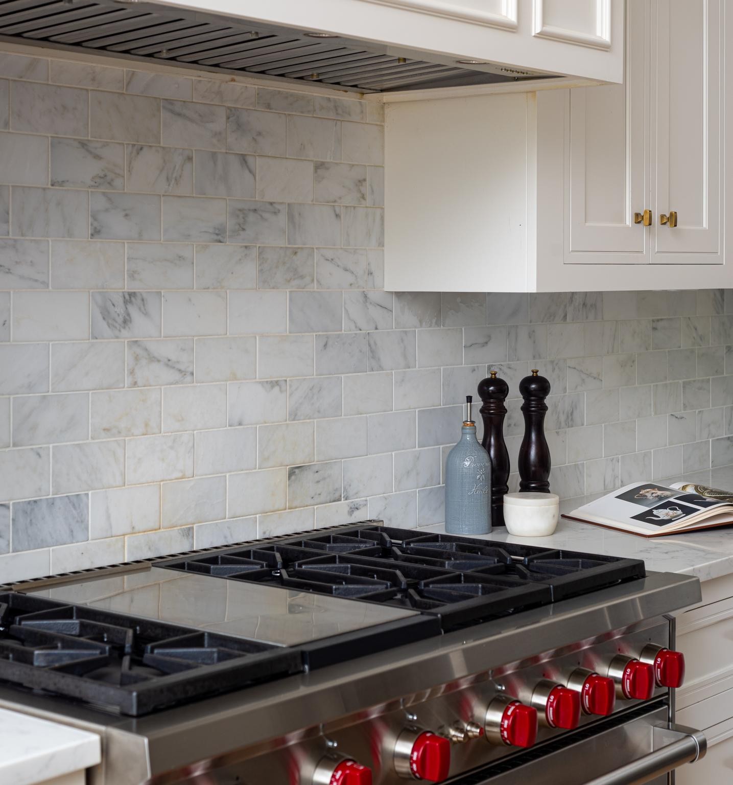 Kitchen range with red knobs, marble backsplash, white cabinetry, and countertop accessories.