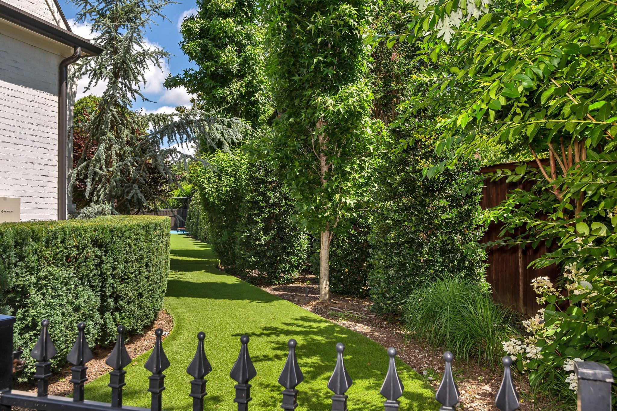 Manicured side yard with a narrow lawn, dense hedges, flowering plants, and black metal fence.