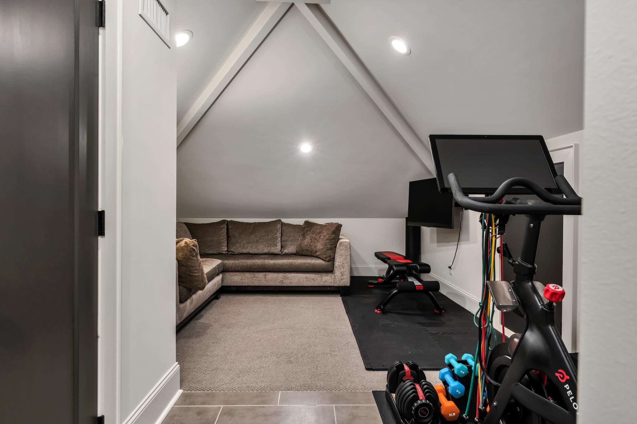 Home gym with a Peloton bike, weights, exercise equipment and a sectional sofa under a sloped ceiling.