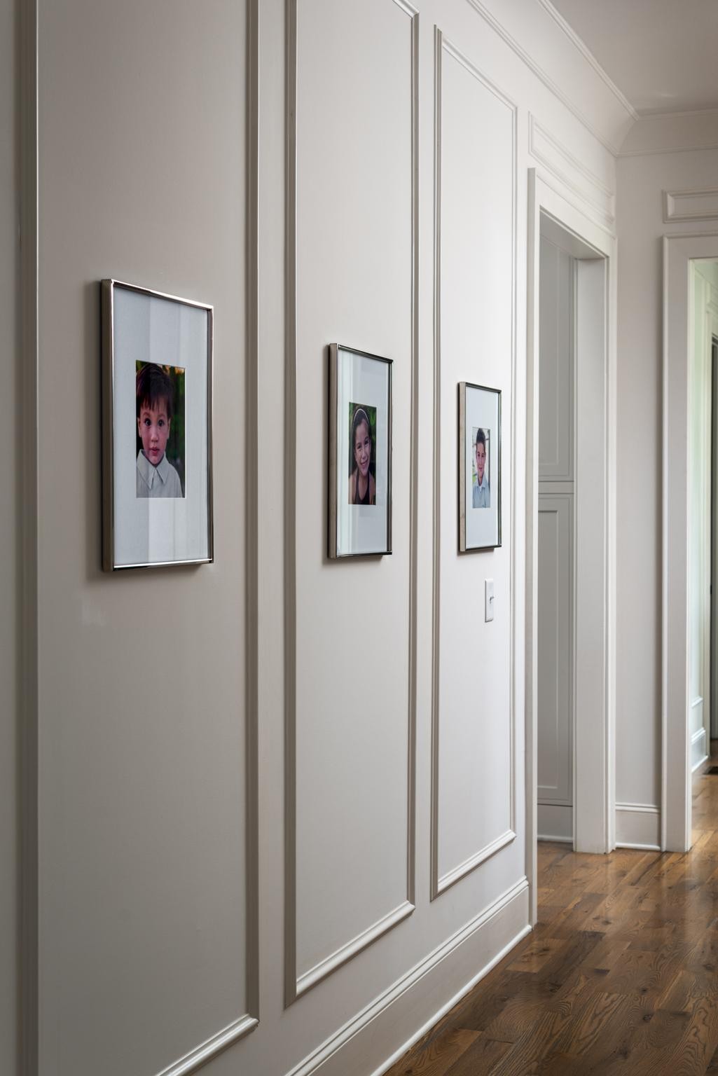Hallway with paneled walls, three framed portraits, and hardwood floors.
