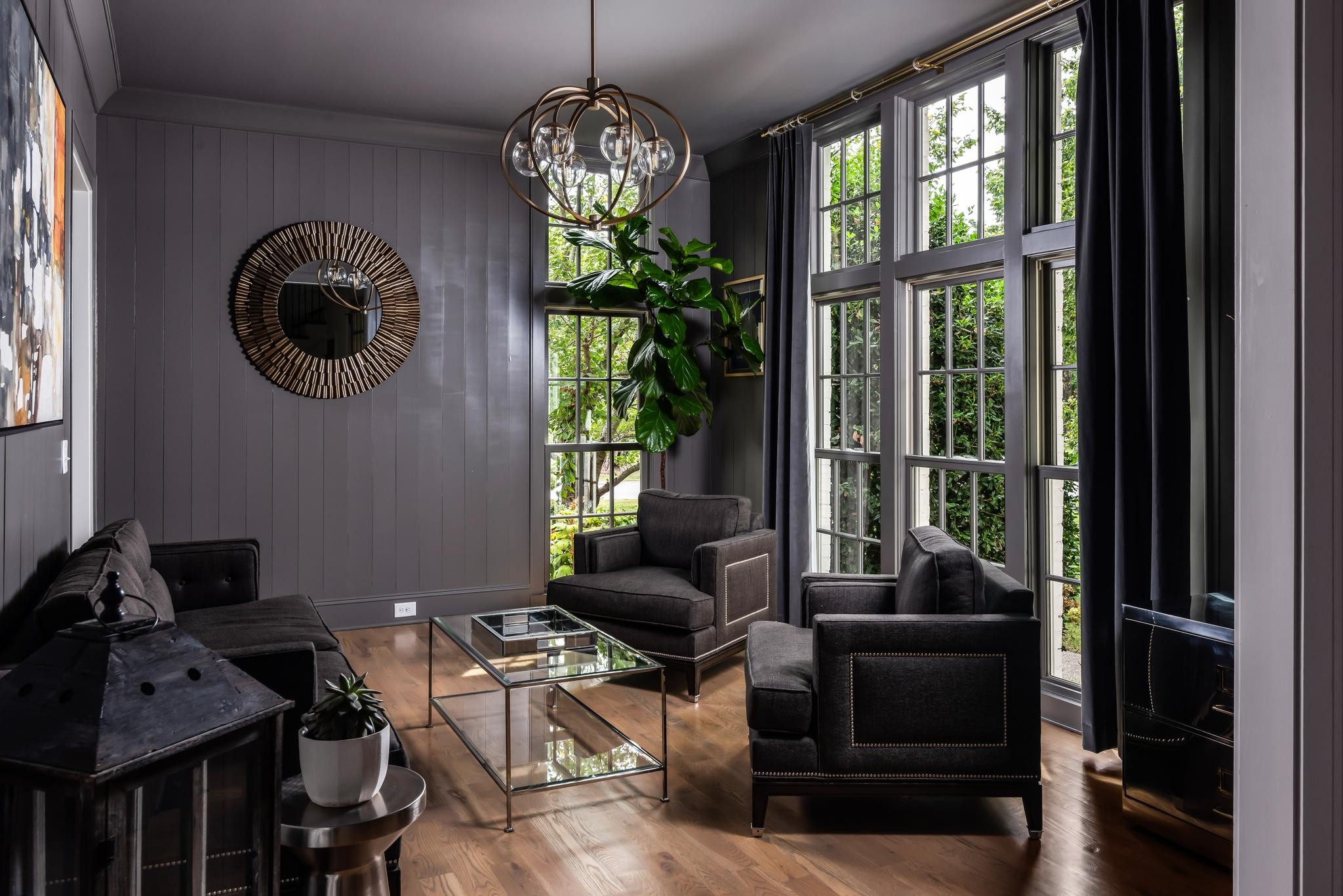 Living room with dark seating, a glass coffee table, and tall windows overlooking greenery.