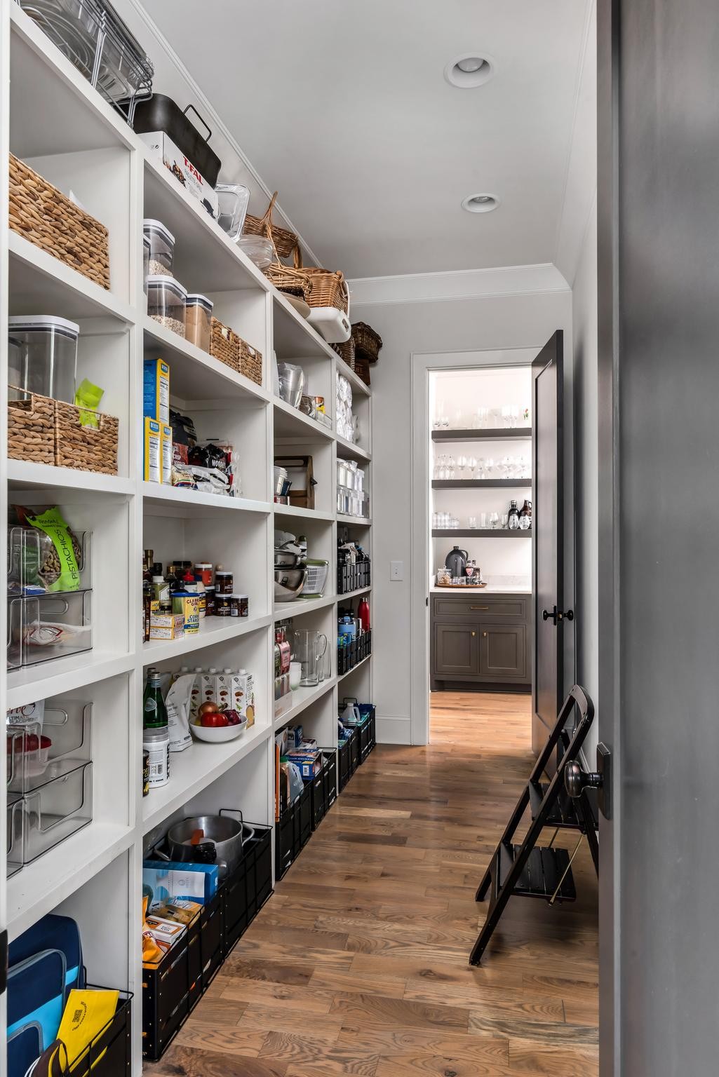 Walk-in pantry with stocked white shelves, woven baskets, black storage bins, and wood flooring.