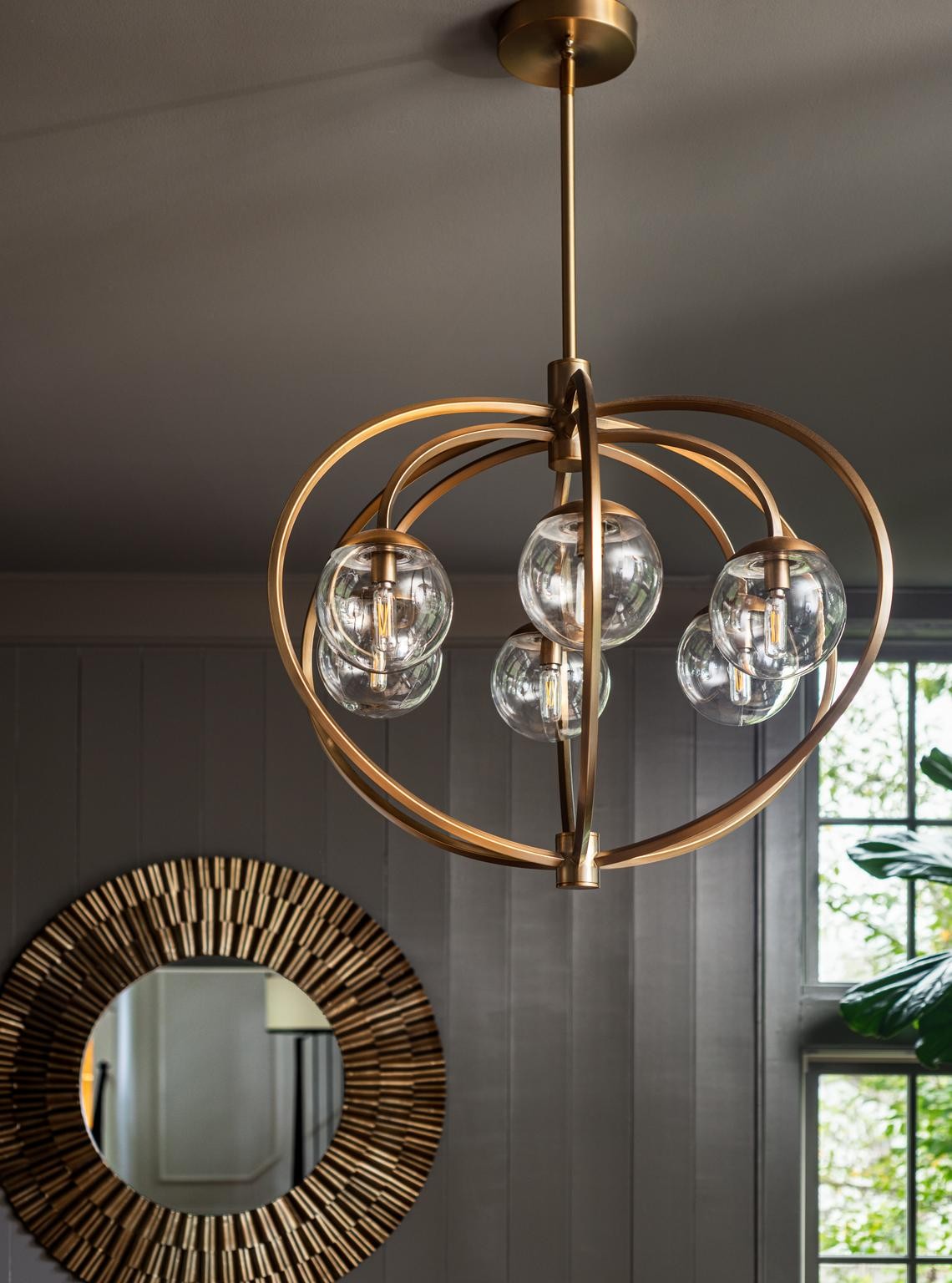 Brass orb chandelier with clear glass globes above a sunburst mirror beside a window.