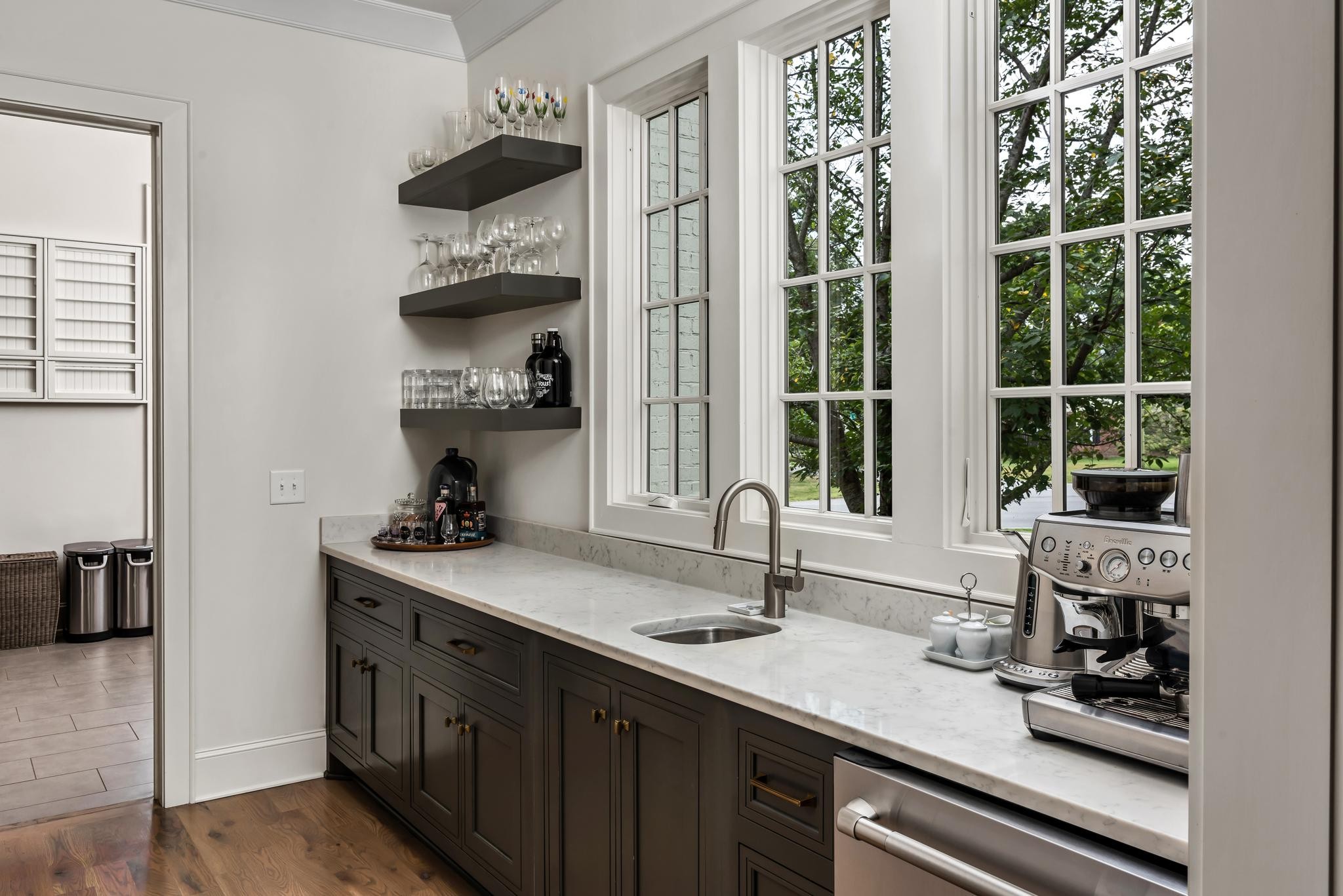 Kitchen beverage station with marble counter, espresso machine, glassware shelves, and large windows.