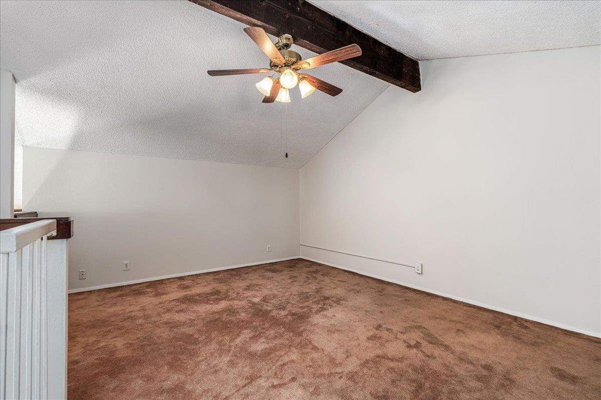 Empty carpeted room with a ceiling fan, exposed beam, and railing.