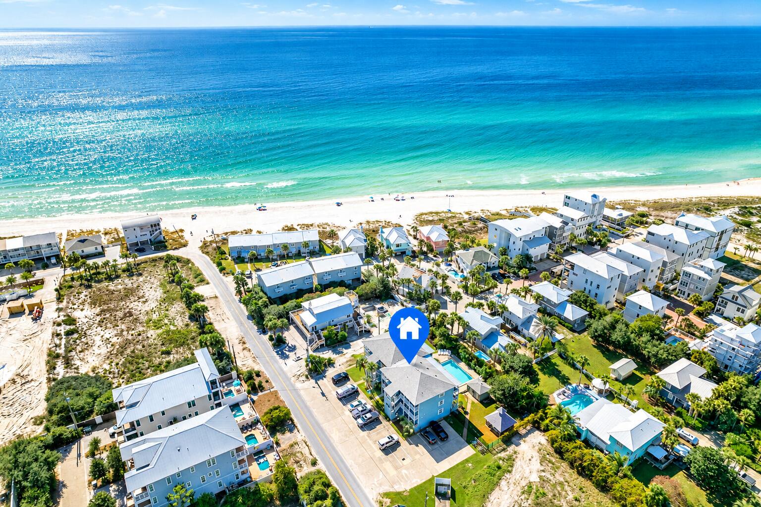 Aerial coastal neighborhood beside turquoise water and a white-sand beach, with homes and pools.