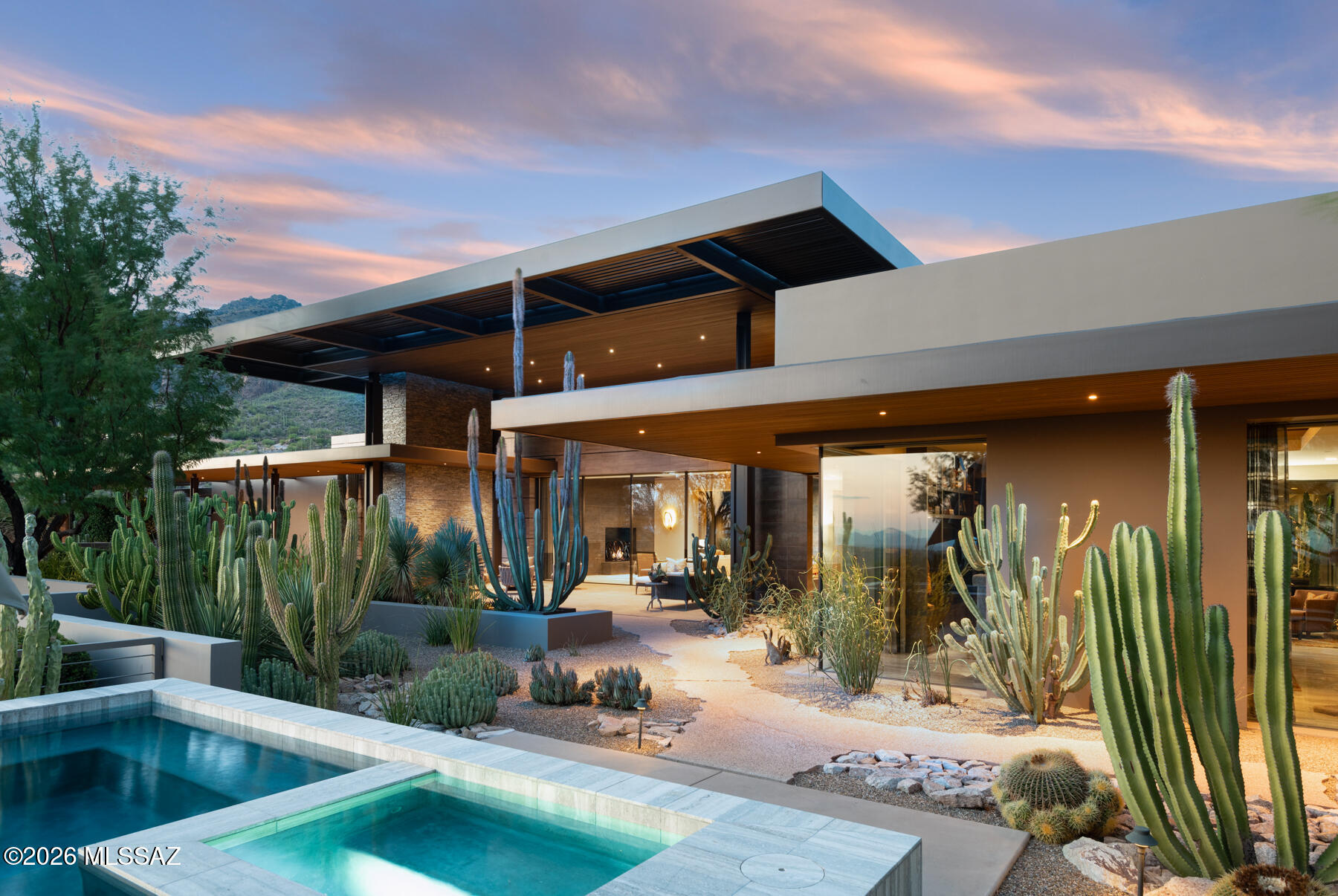 Modern desert home with angular roof, cactus landscaping, and illuminated pool at dusk.