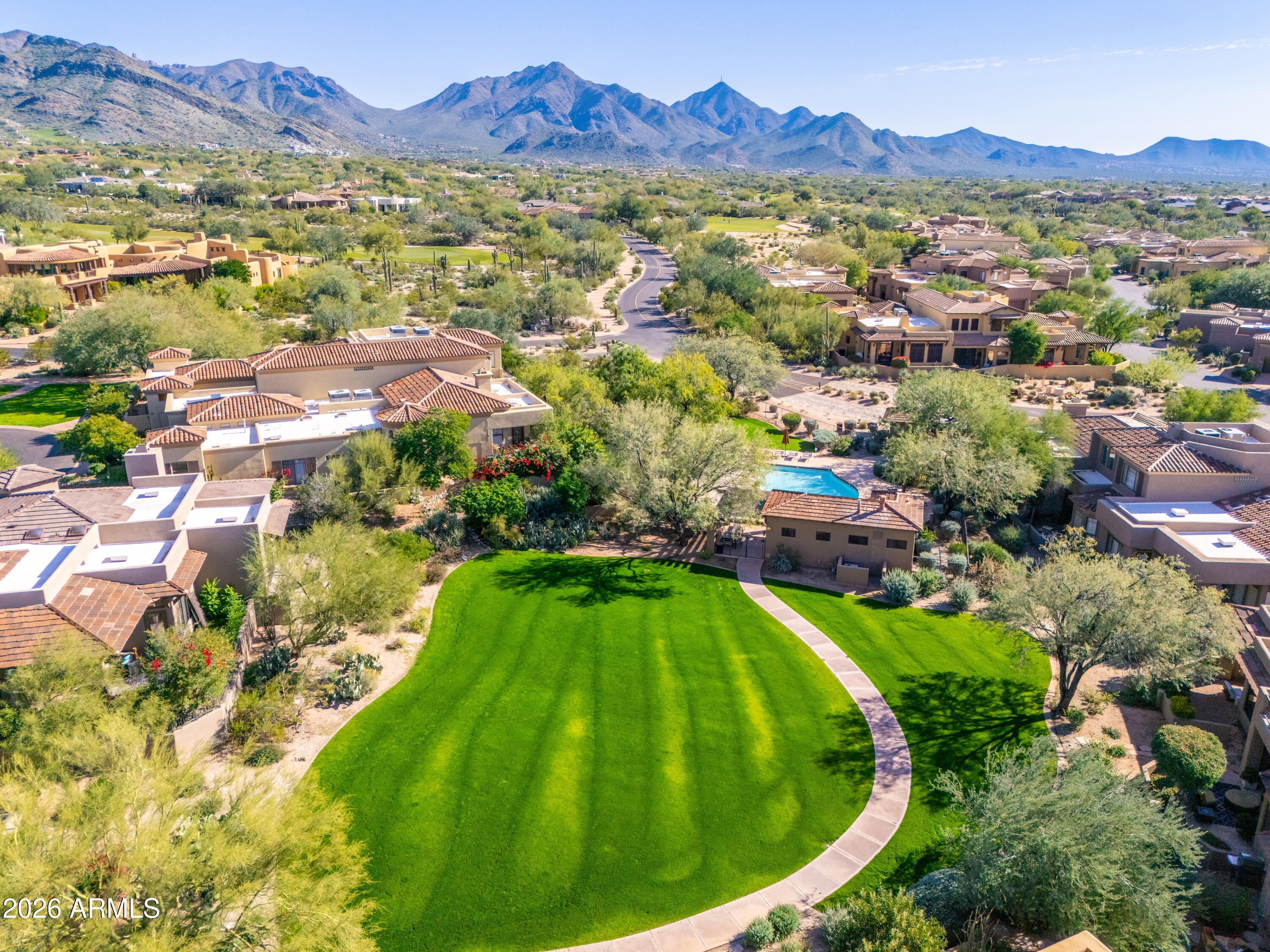 Aerial view of homes around a broad green lawn, with a pool and mountain ranges beyond.