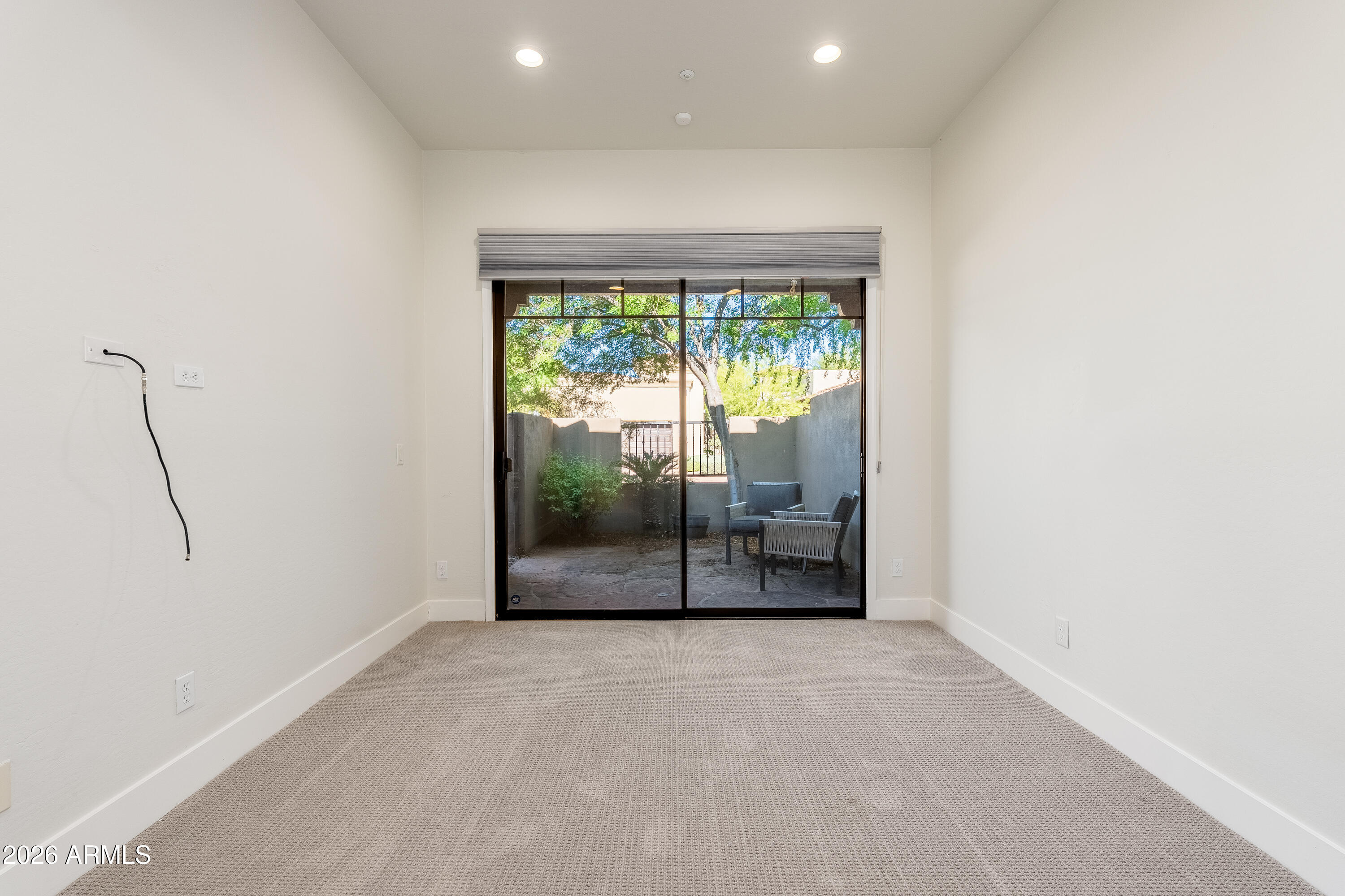 Empty bedroom with carpet and sliding glass doors opening to a walled patio with chairs and greenery.