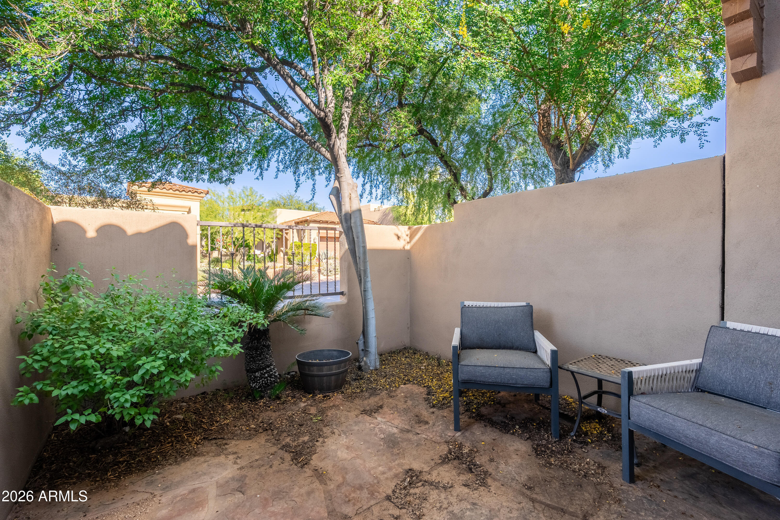 Walled patio with gray seating, leafy shrubs, and mature trees providing shade.