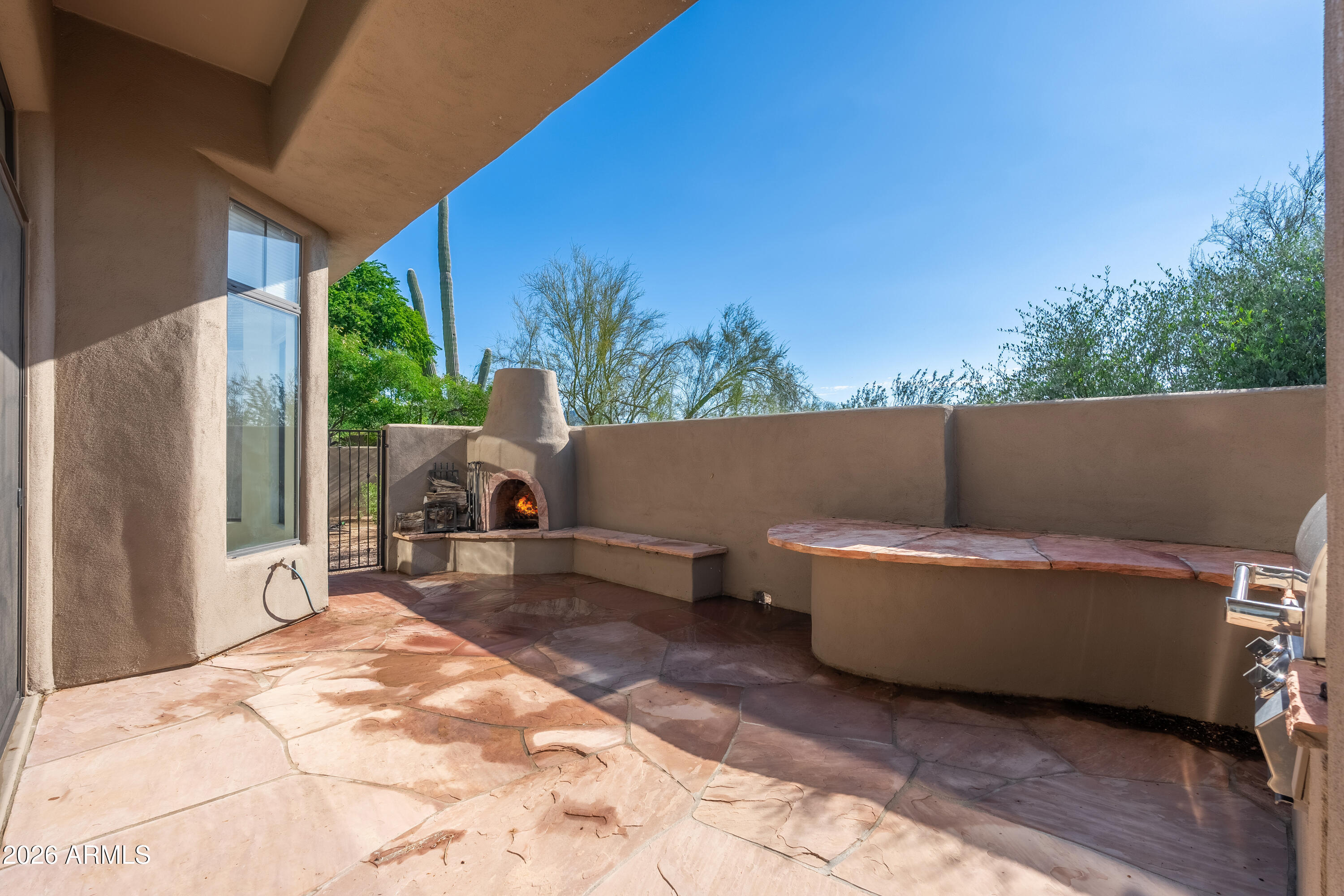Covered flagstone patio with a built-in fireplace and bench seating, overlooking desert trees and saguaro cacti.