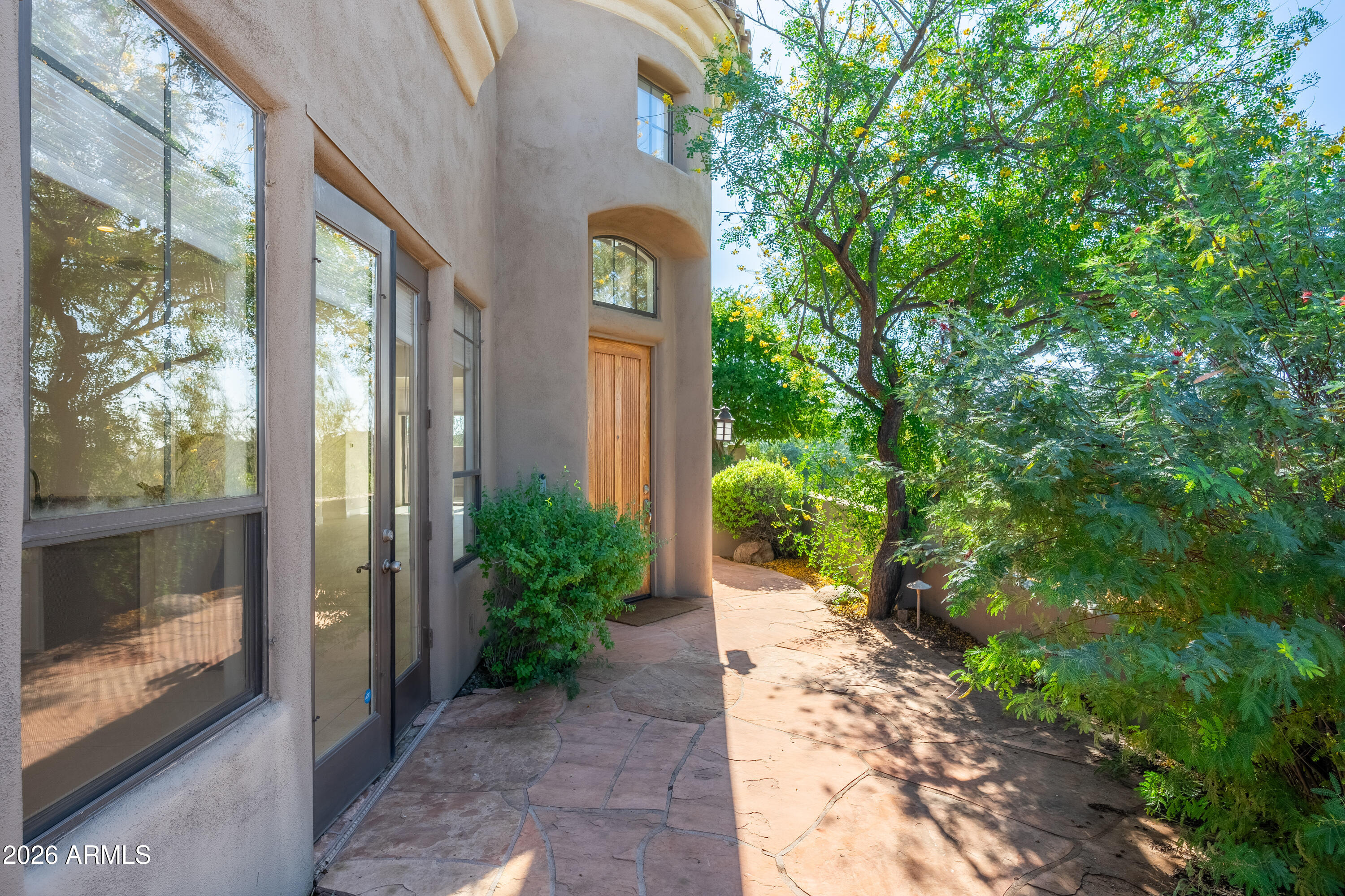 Stucco home entrance with an arched doorway, wooden door, and shaded stone walkway.