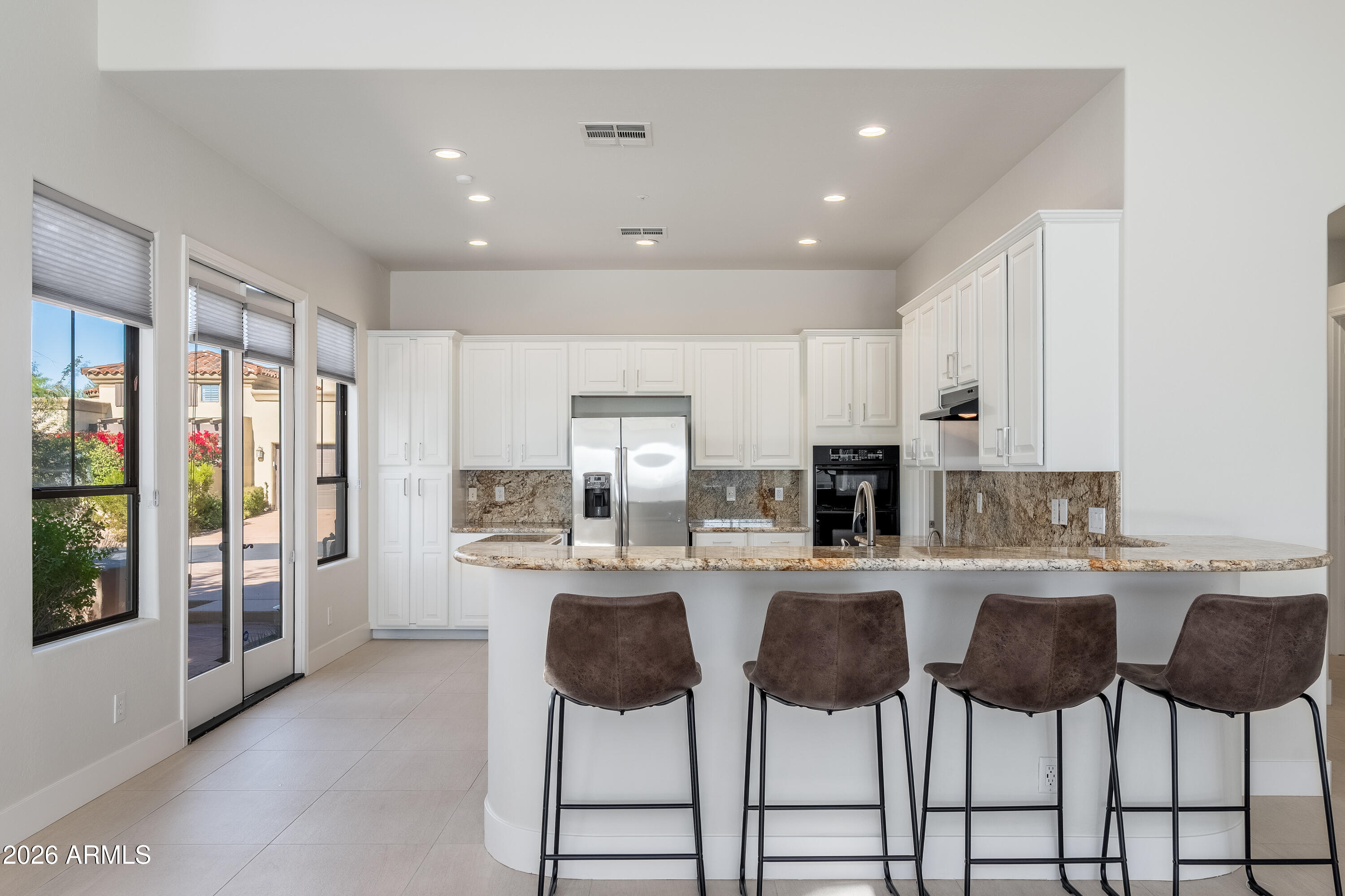 Bright kitchen with white cabinetry, stone counters, stainless steel appliances, and a curved breakfast bar with four stools.