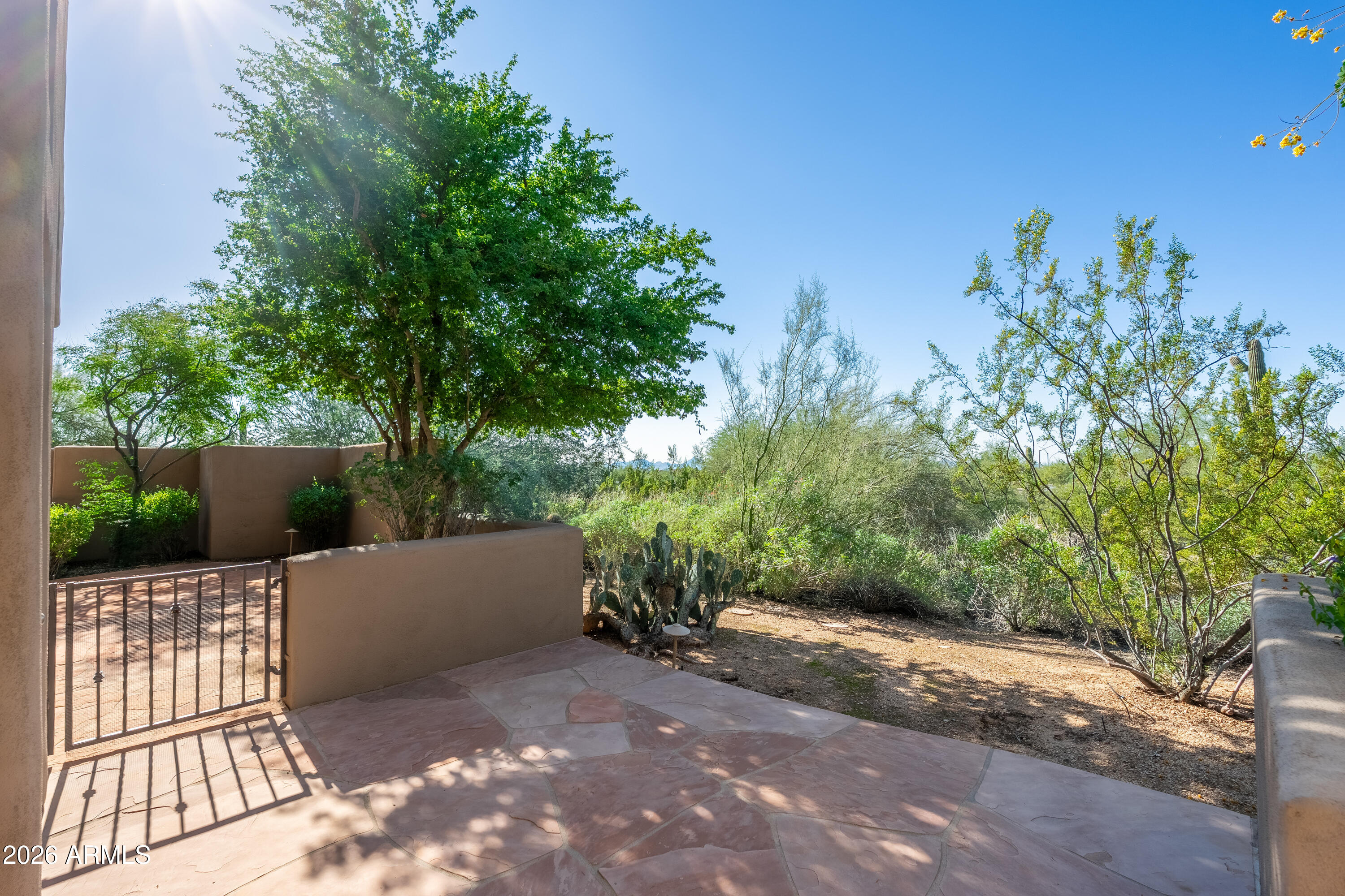 Stone patio with a metal gate, stucco walls, mature trees, and desert landscaping.