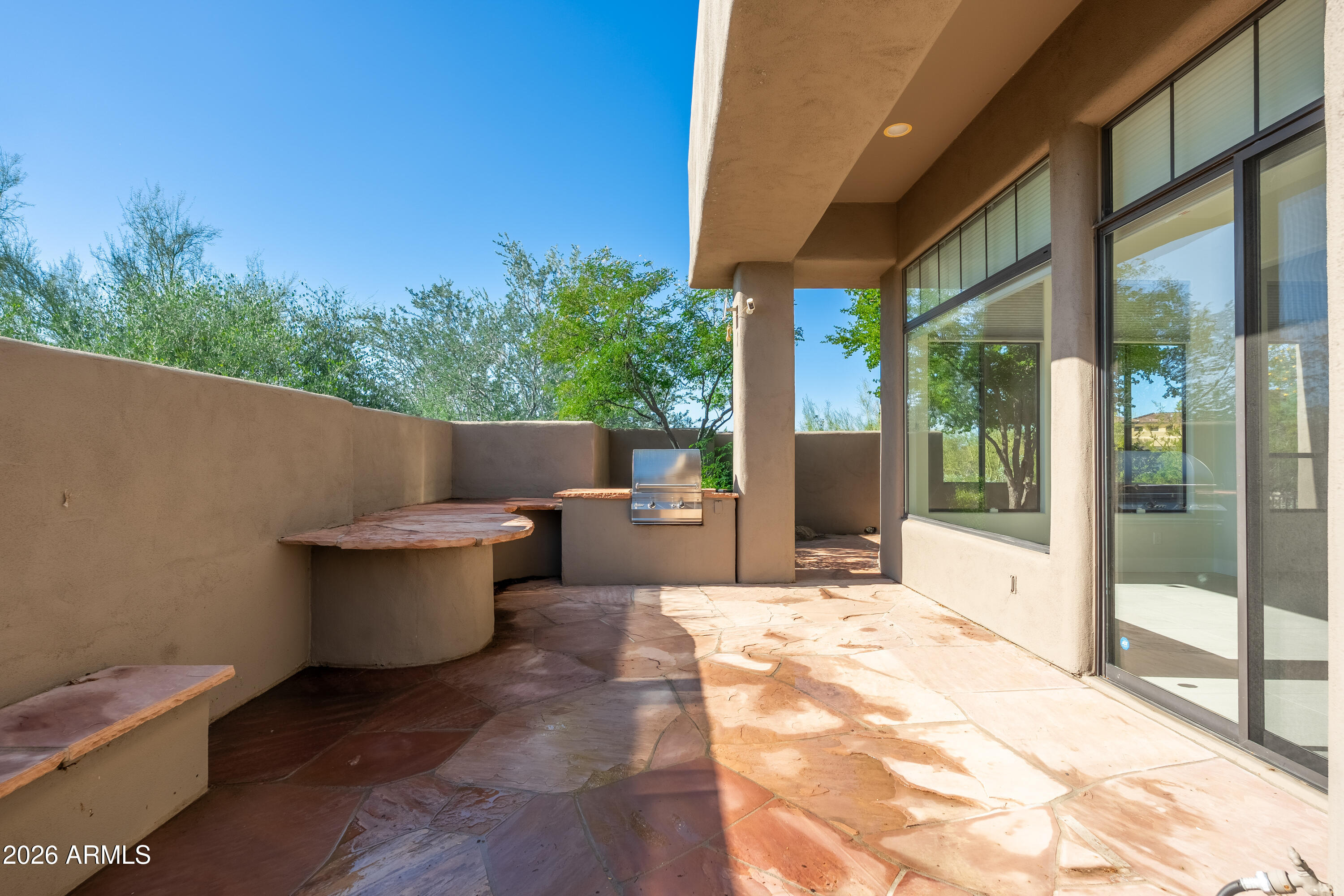 Covered stone patio with a built-in grill, curved seating, and large windows.