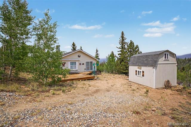 Small house with a wooden deck and detached shed in a wooded gravel clearing.