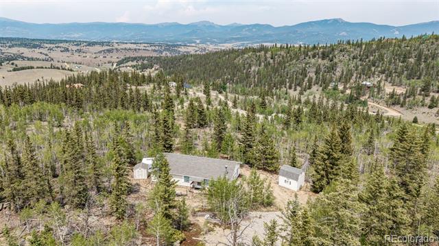 Aerial view of a home and outbuildings among pine trees, with mountain ranges in the distance.