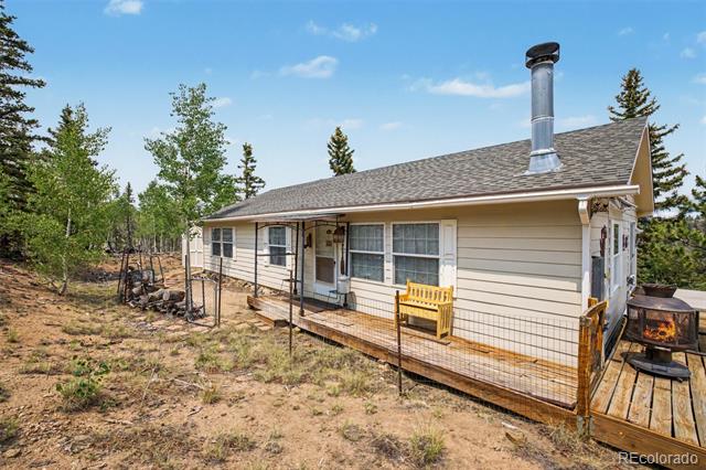 Single-story beige house with a raised wooden deck, yellow bench, and metal chimney in a wooded yard.
