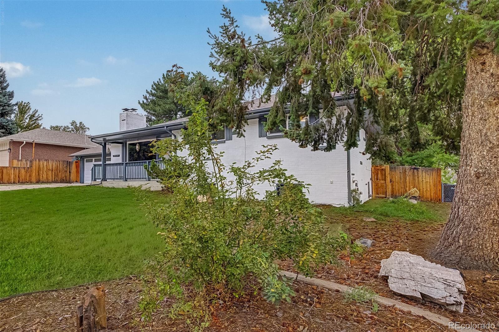 White brick house beside a broad lawn, with a covered porch, wood fencing, and mature trees.