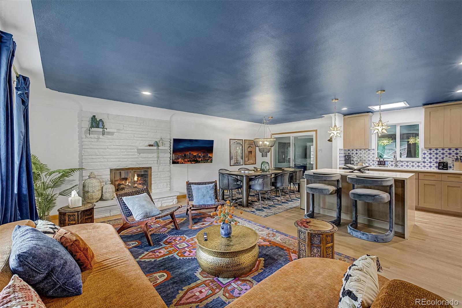 Open living room with a white stone fireplace, deep blue ceiling, and adjoining dining area and kitchen.