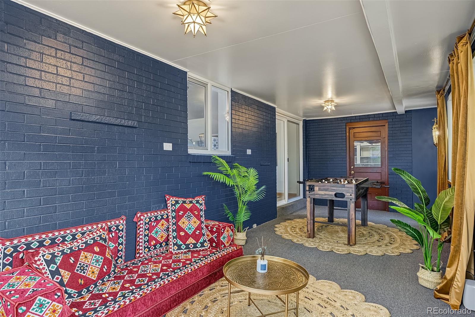Furnished sunroom with navy brick walls, a patterned red sofa, potted plants, and a foosball table.