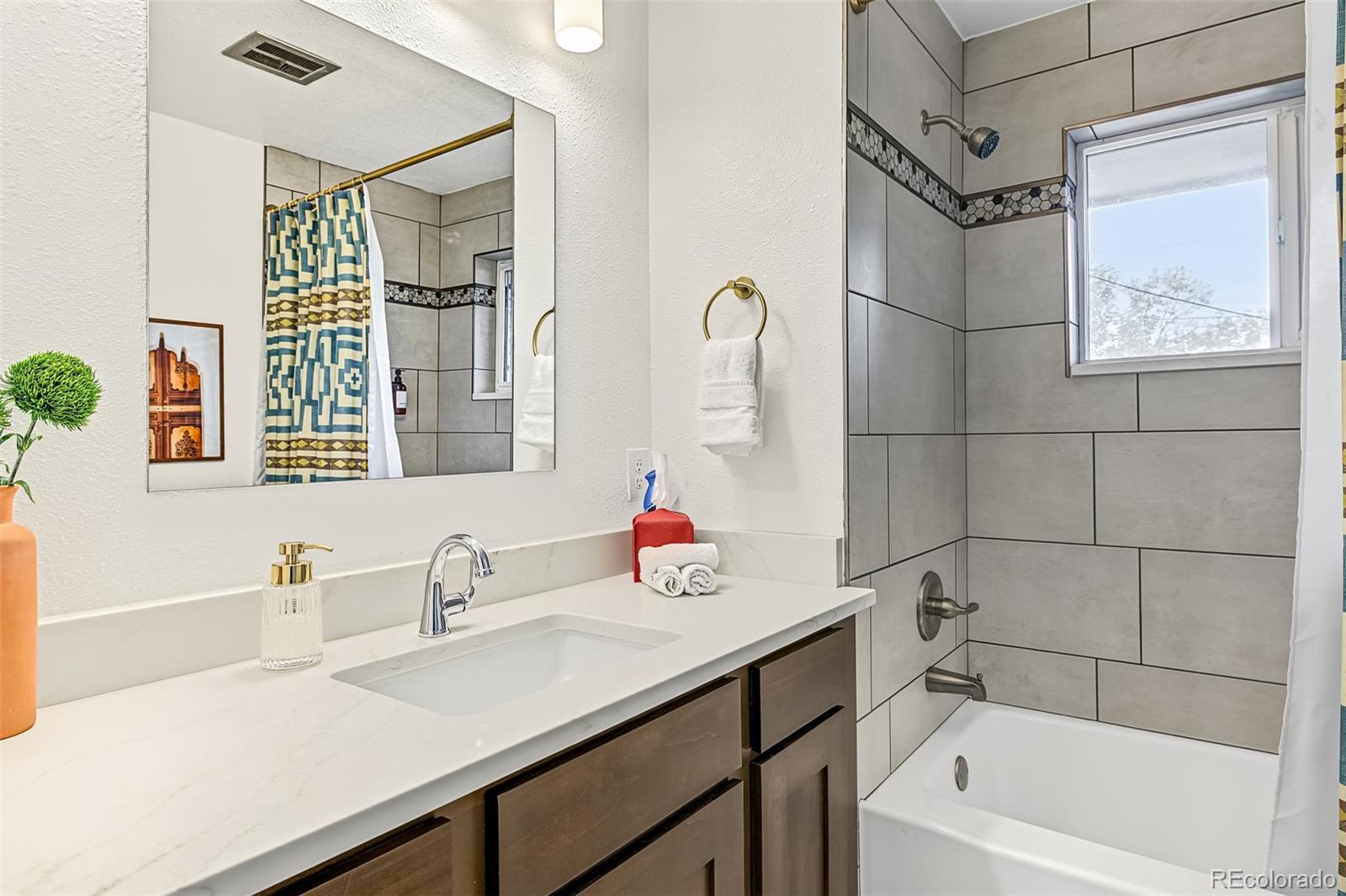 Updated bathroom with a white vanity and sink beside a gray-tiled tub and window.