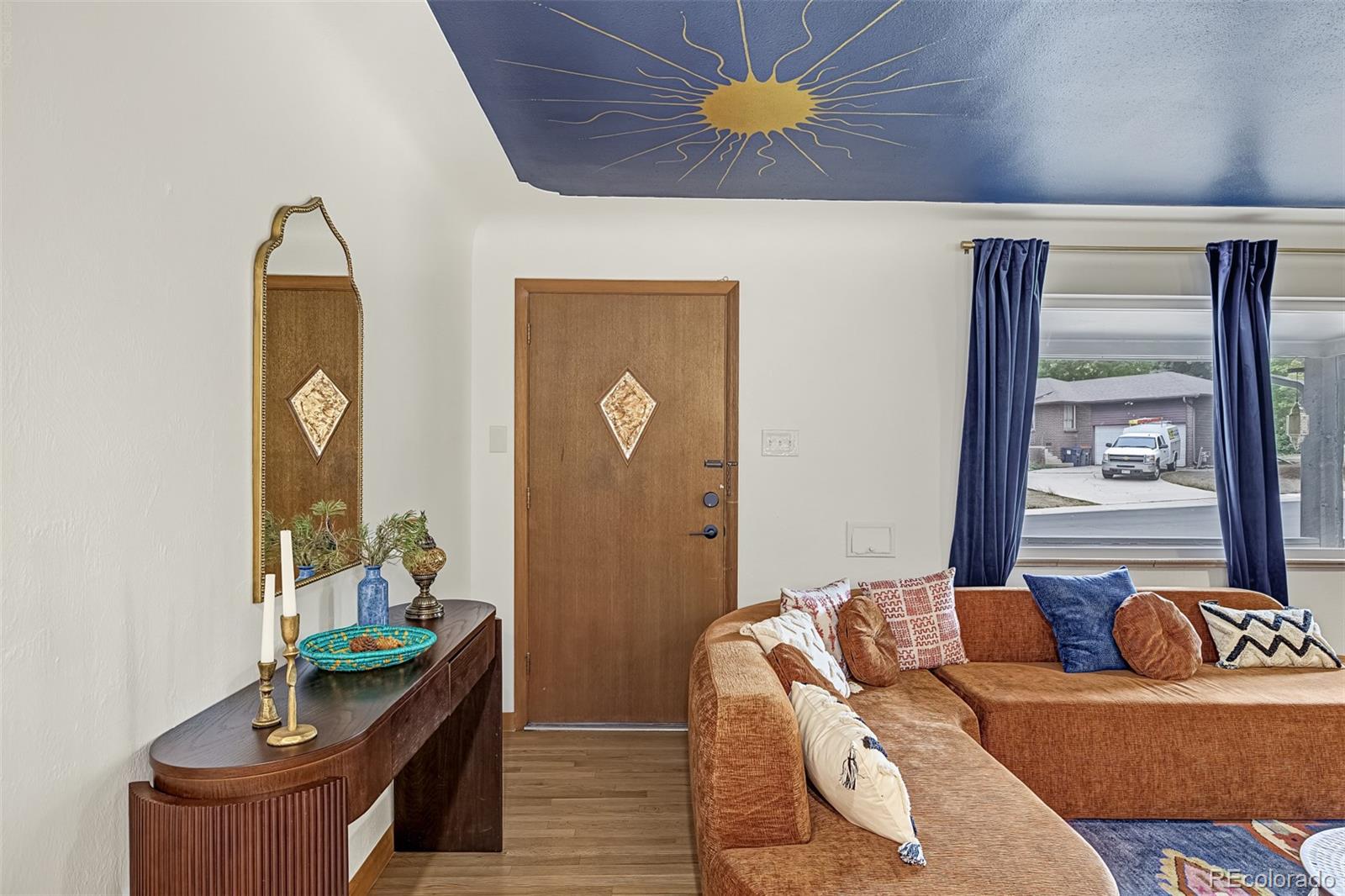 Living room with an orange sectional, wooden entry door, and blue sunburst ceiling.