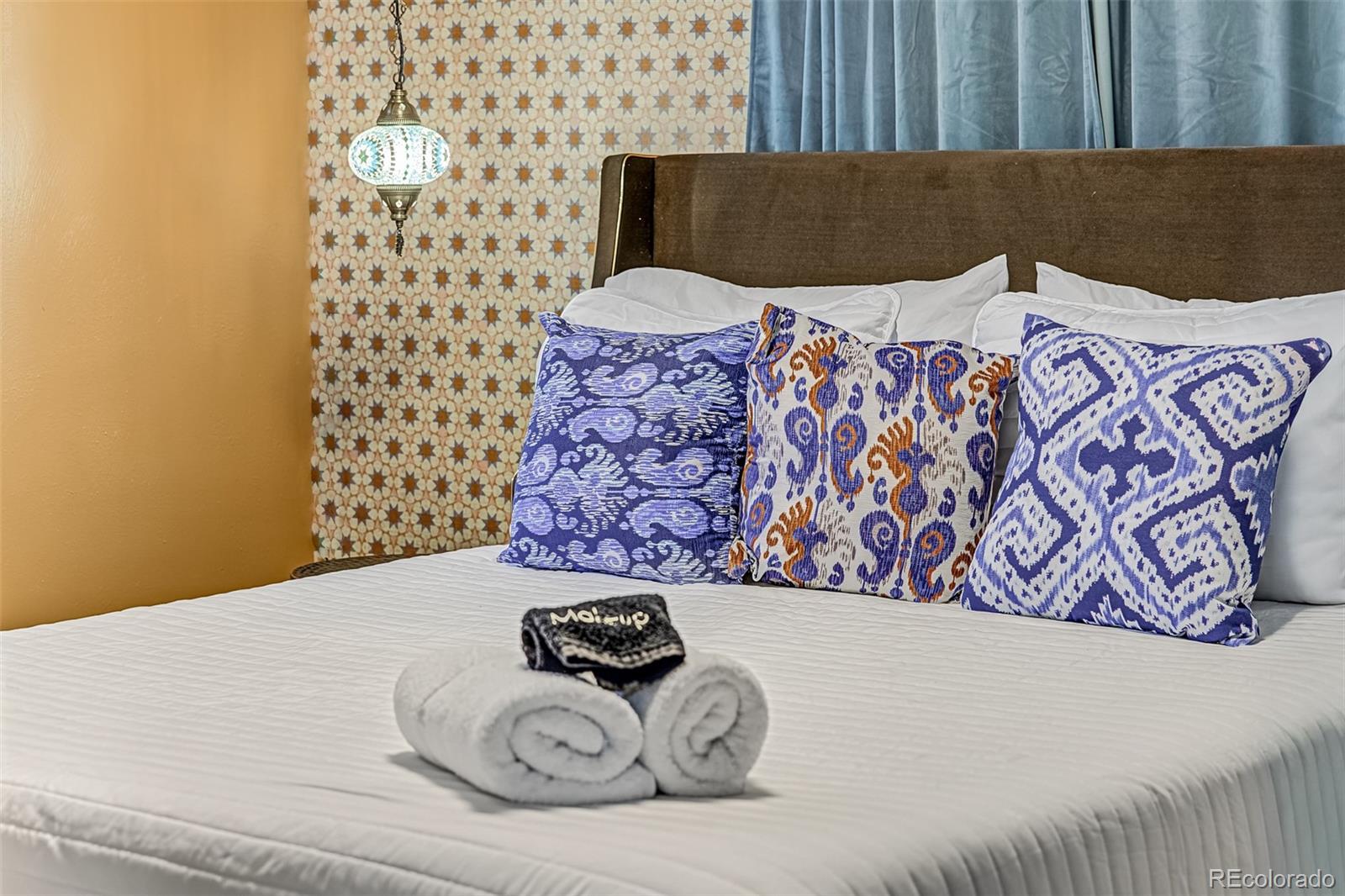 Bedroom with patterned accent wall, blue curtains, decorative pillows, and rolled towels on the bed.