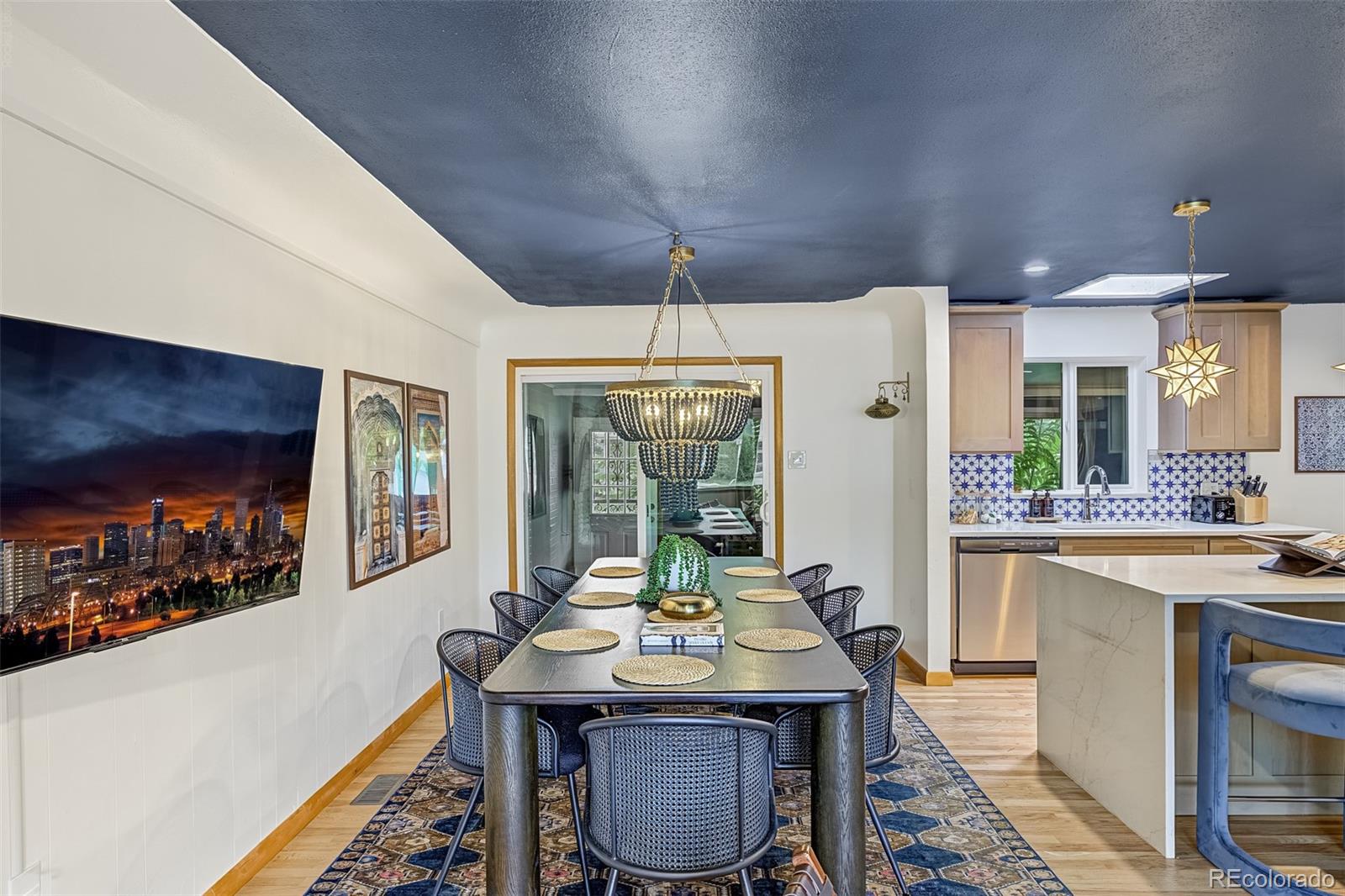 Dining room with a long table beneath a tiered chandelier, opening onto a kitchen with patterned tile.