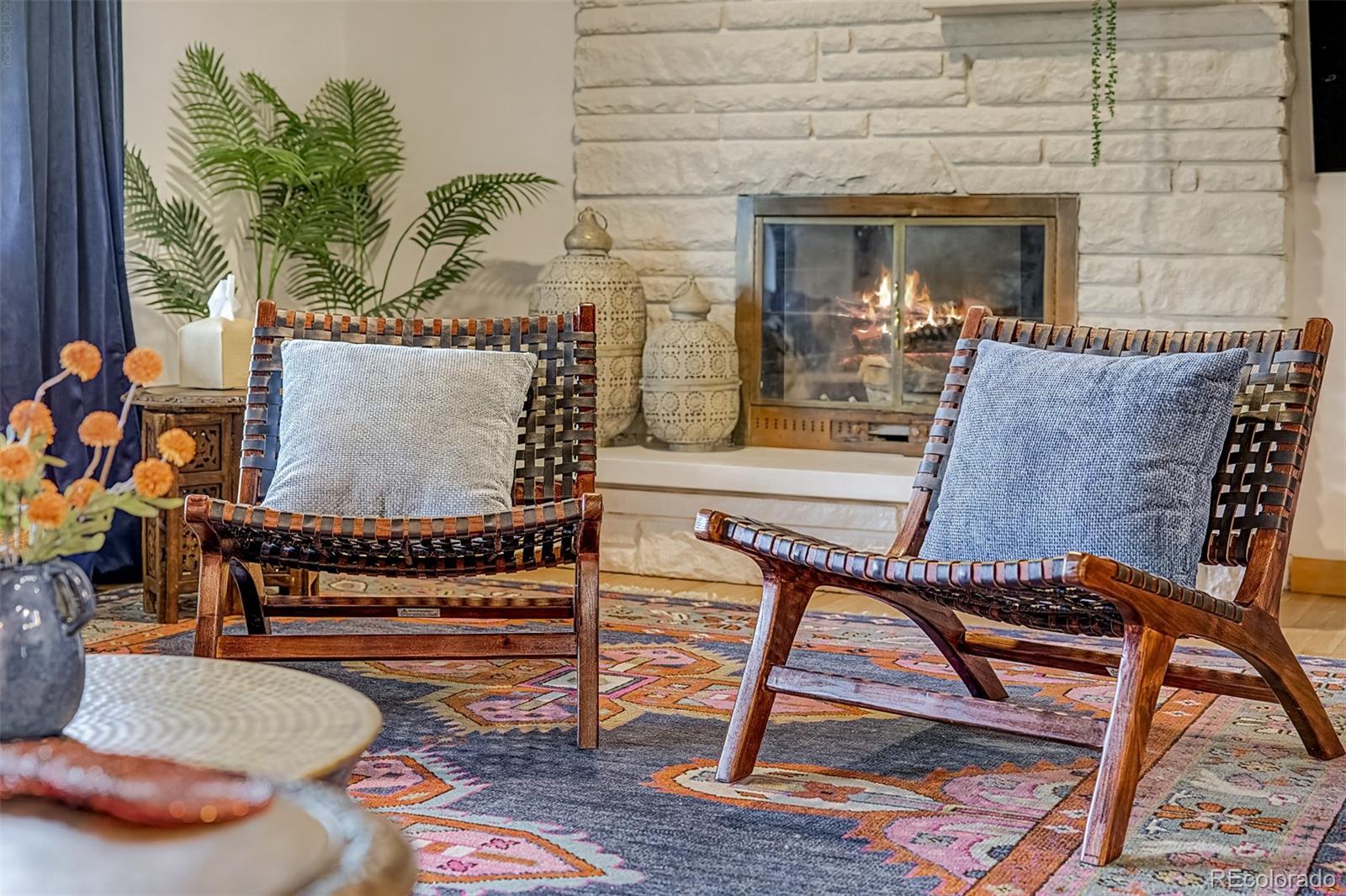 Living room with woven lounge chairs, a lit stone fireplace, and a colorful patterned rug.