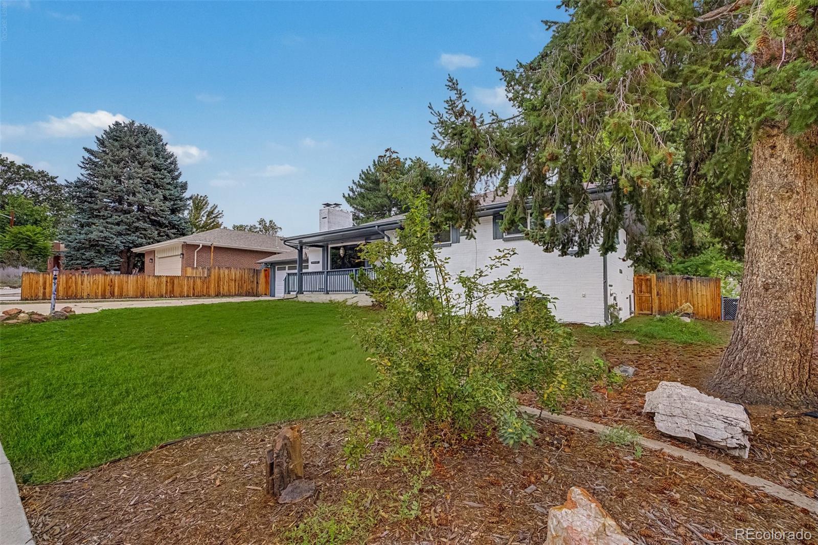 White brick home with covered porch, green lawn, wooden fencing, and mature trees in the backyard.