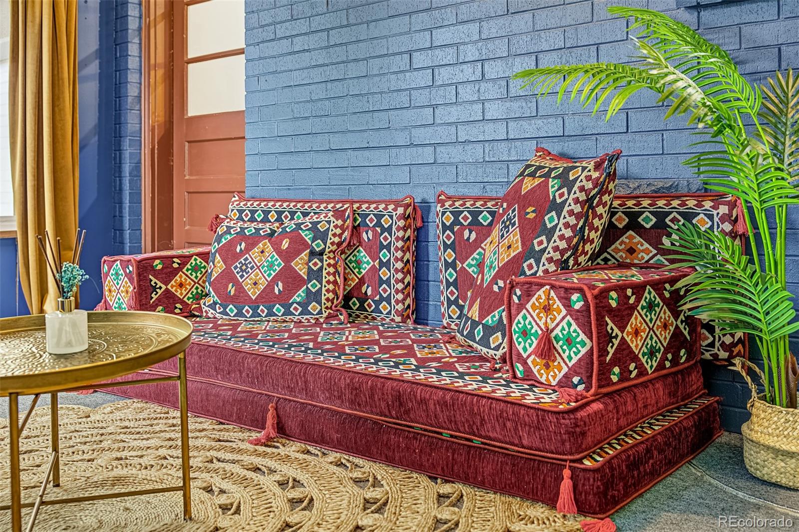 Patterned burgundy sofa and cushions against a blue brick wall, with a brass table and potted palm.