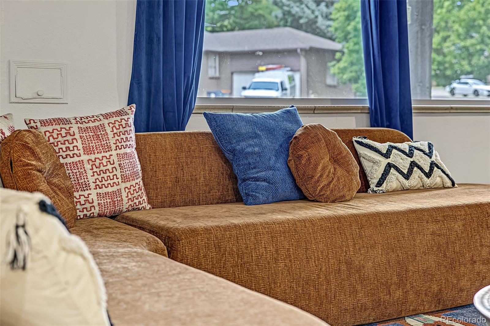 Brown sectional sofa with patterned cushions sits beneath blue curtains and a large window.