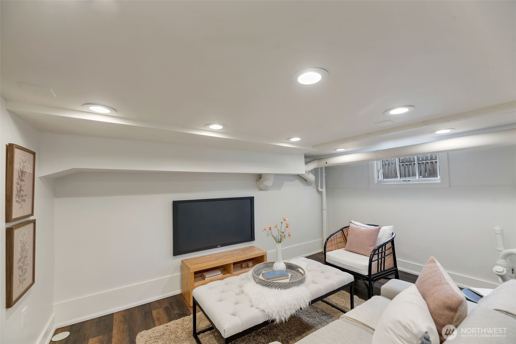 Basement living room with a wall-mounted TV, recessed lights, and white tufted ottoman.