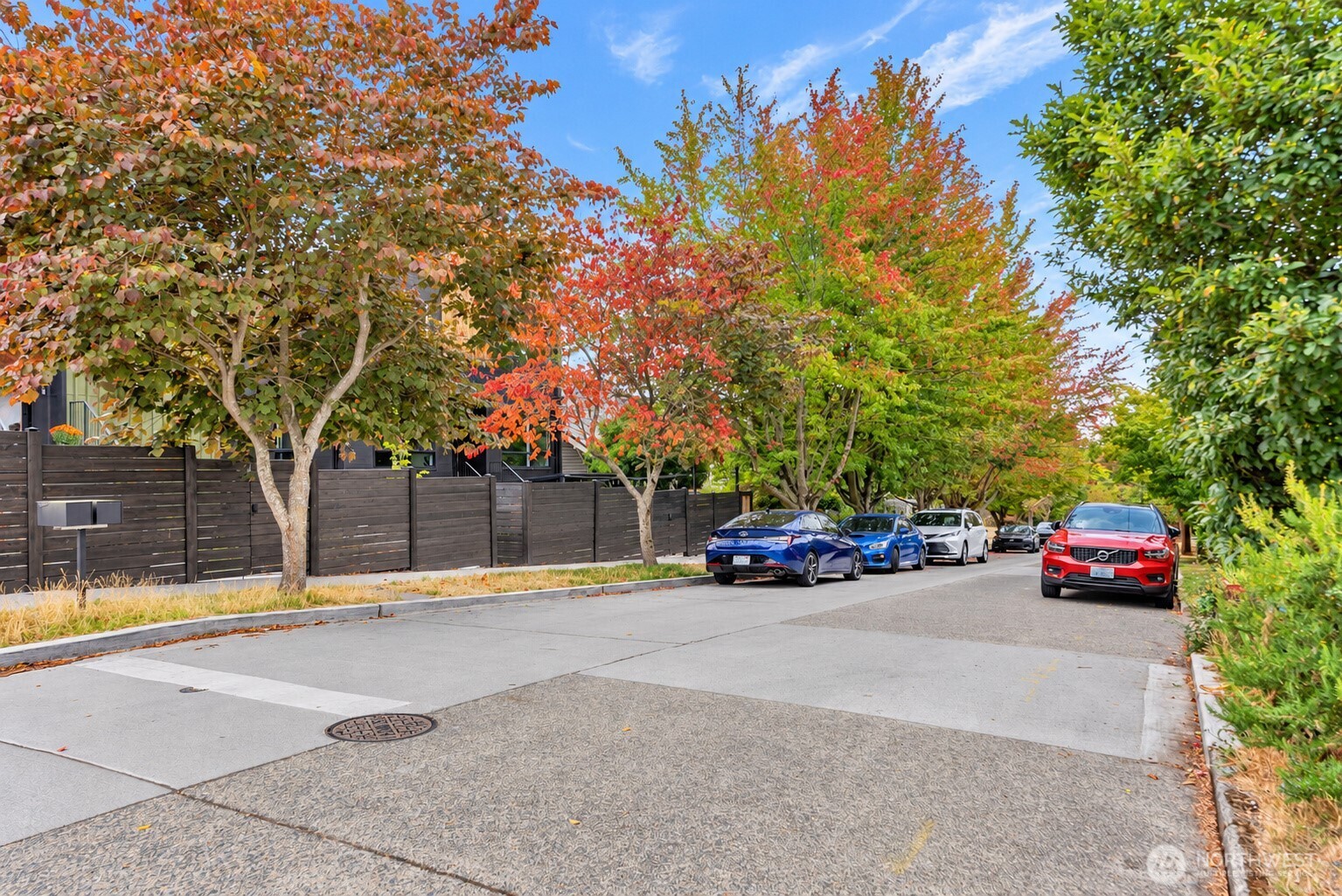 Residential street lined with autumn trees, parked cars, and a dark fence partly screening the property.