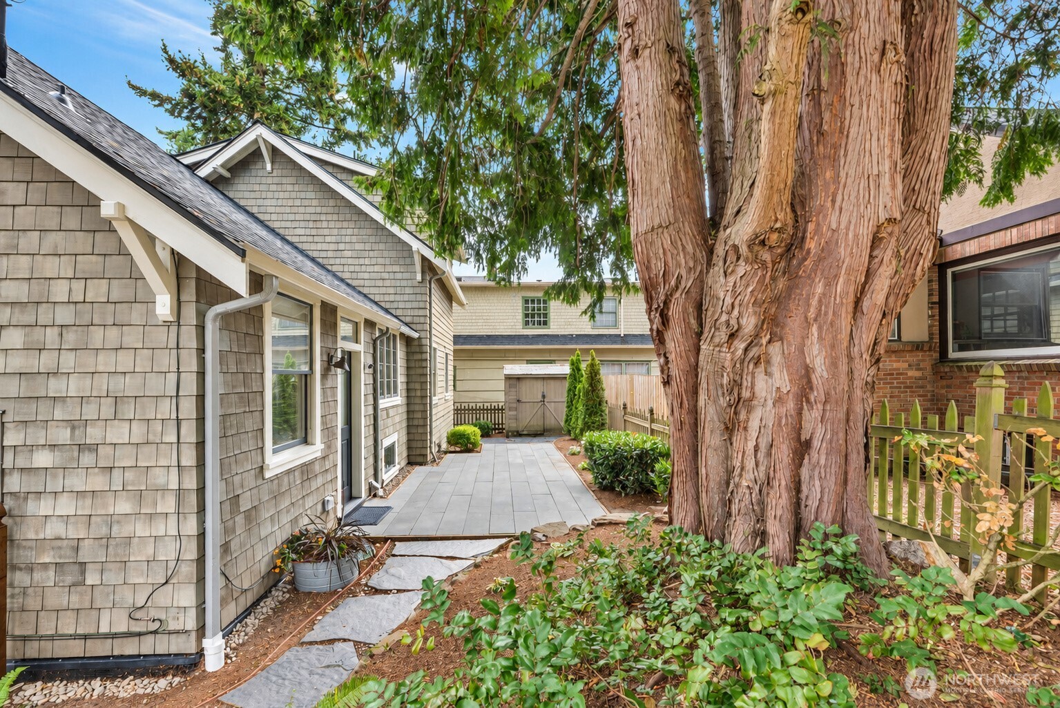 Backyard patio beside a gray shingle-sided house, shaded by a large mature tree.