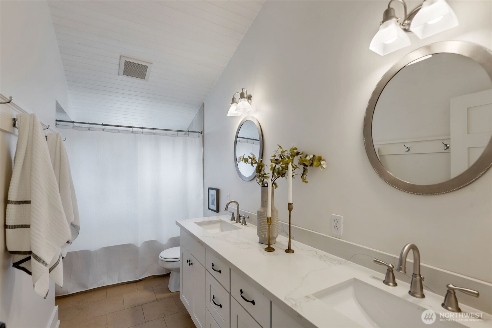 Bathroom with a double-sink vanity, round mirrors, and a curtained bathtub beneath a sloped ceiling.