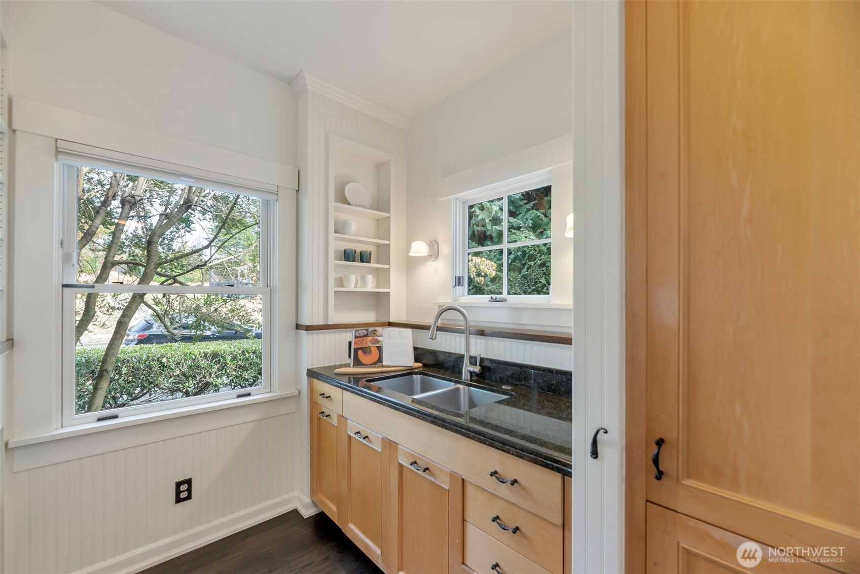 Bright kitchen with light wood cabinetry, dark stone counters, and a double-basin sink beside large windows.