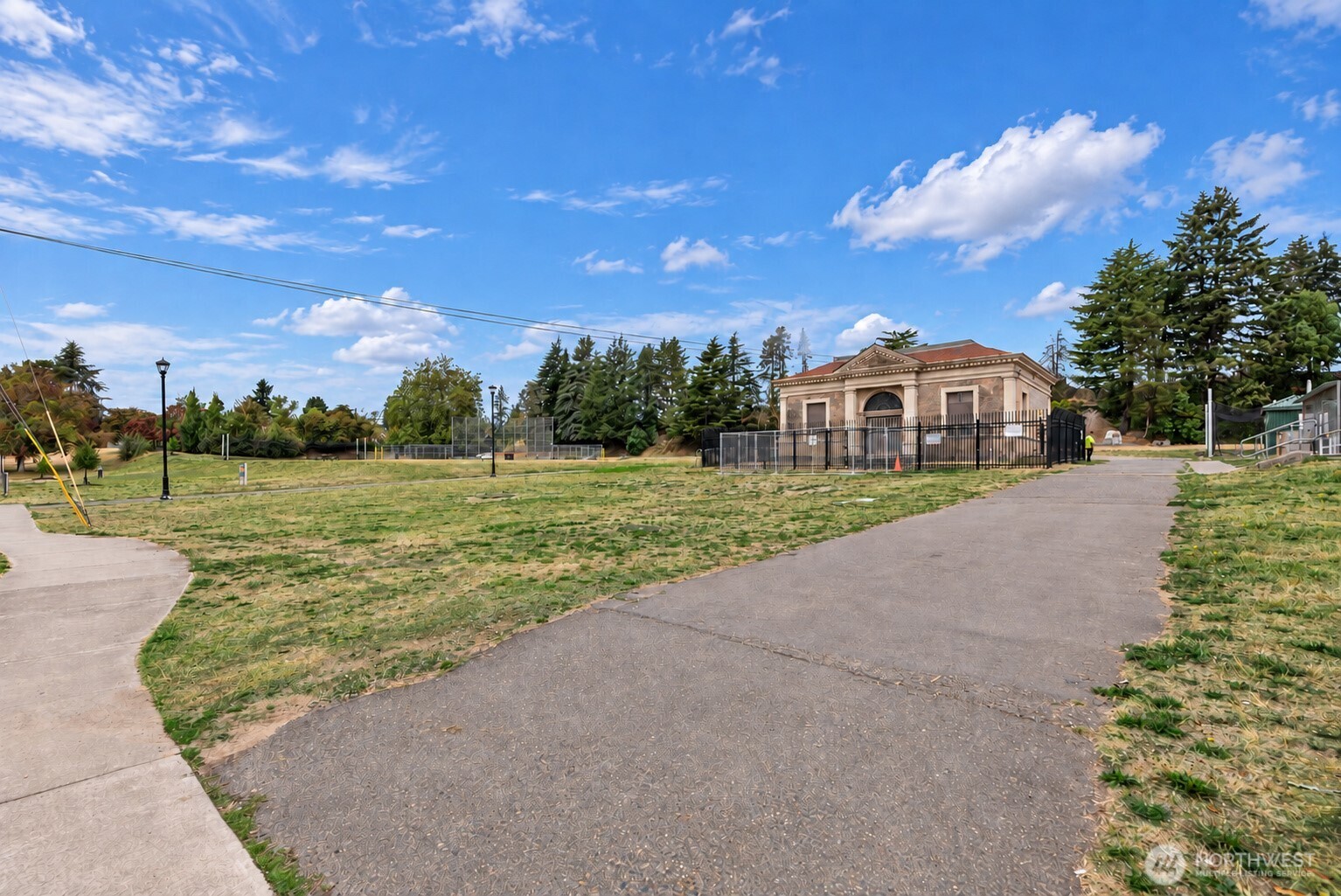 Park path leading across grass to a fenced classical-style building beneath a blue sky.
