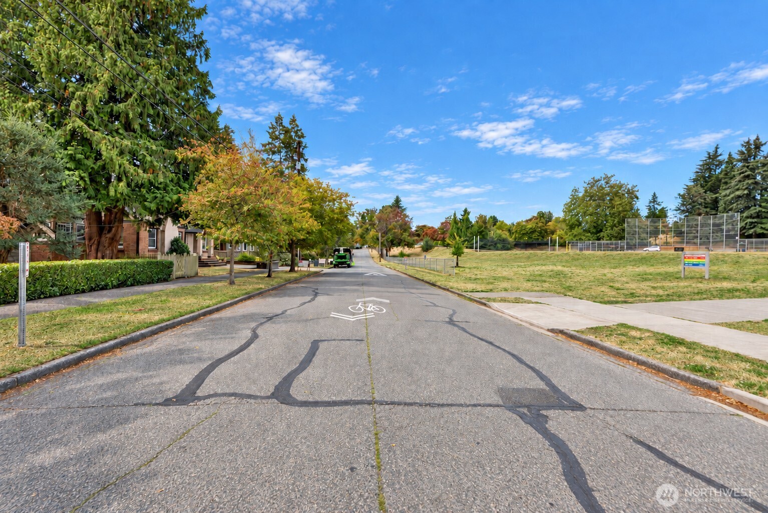 Residential street with trees and homes beside a fenced grassy lot and recreation courts.