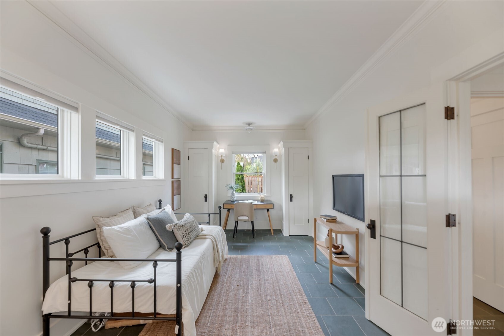 Staged bedroom with a black metal daybed, desk, television, and blue-gray tile floor.