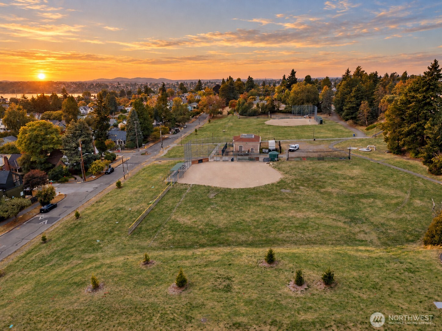 Aerial view of a neighborhood park with a baseball diamond and fenced courts at sunset.