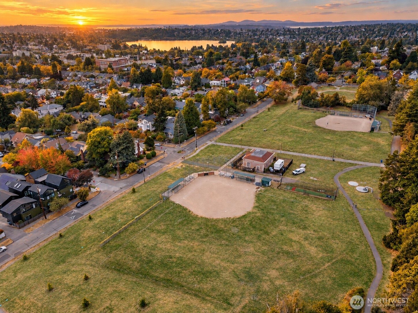 Aerial view of a Seattle neighborhood and park with baseball fields, a lake, and mountains at sunset.