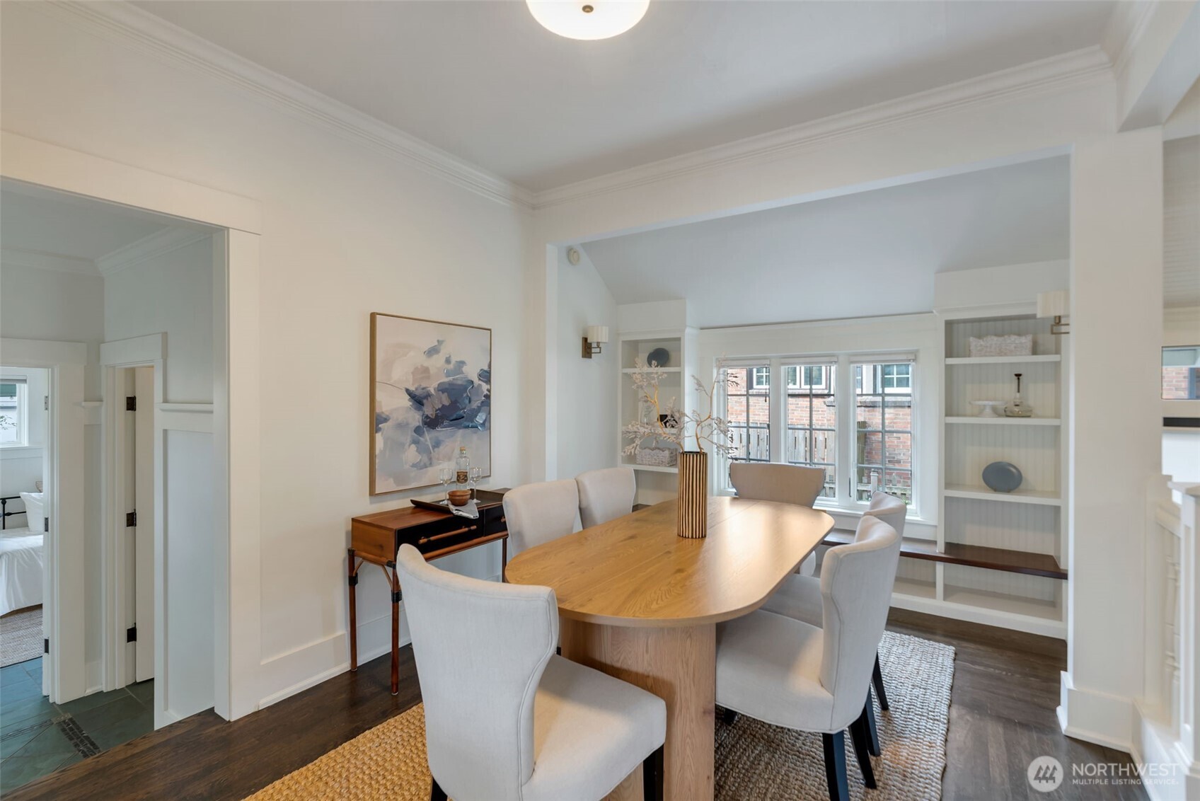 Dining room with an oval wood table, upholstered chairs, and built-in shelving beside large windows.