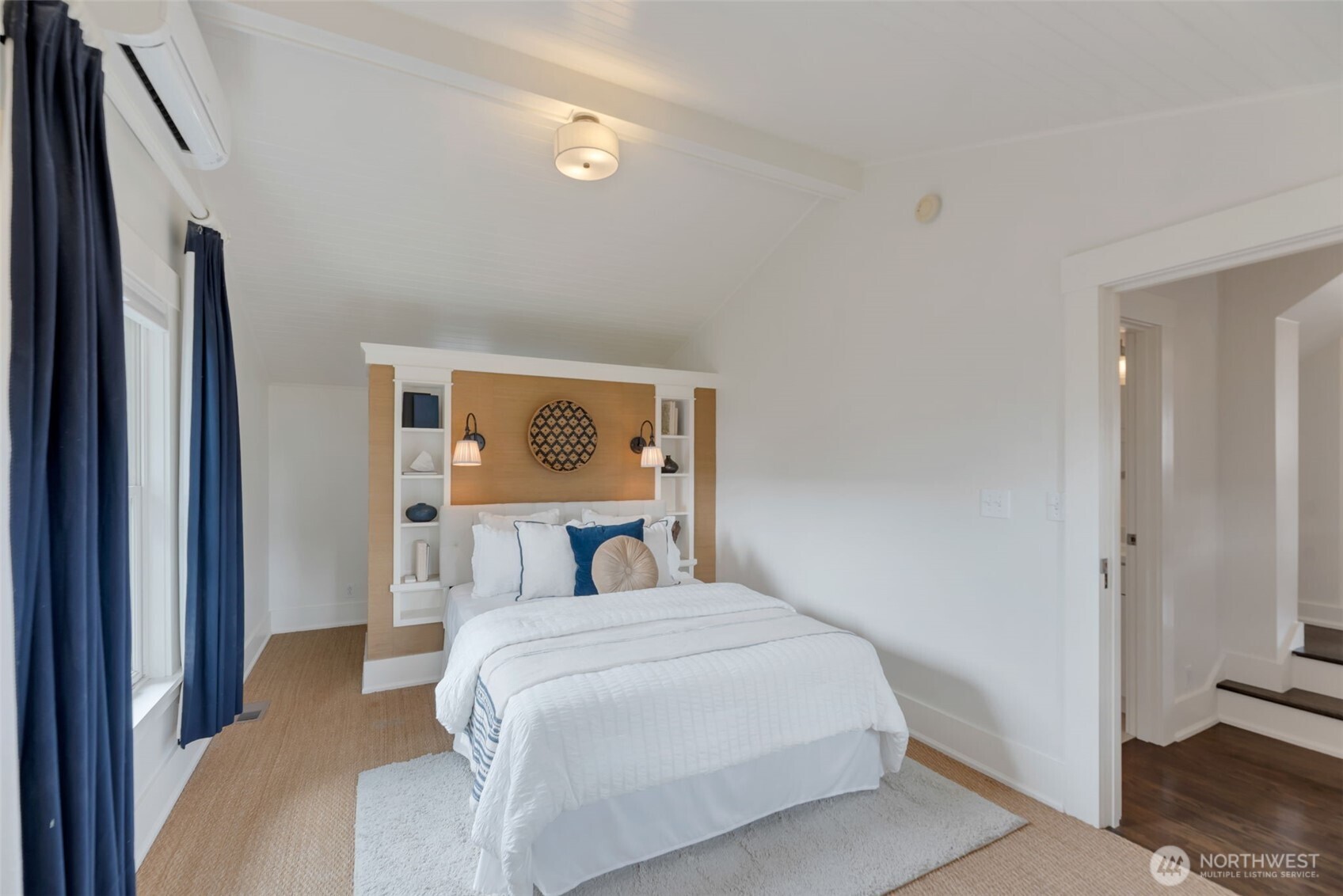 Staged bedroom with a white bed, built-in wood headboard, navy curtains, and vaulted ceiling.